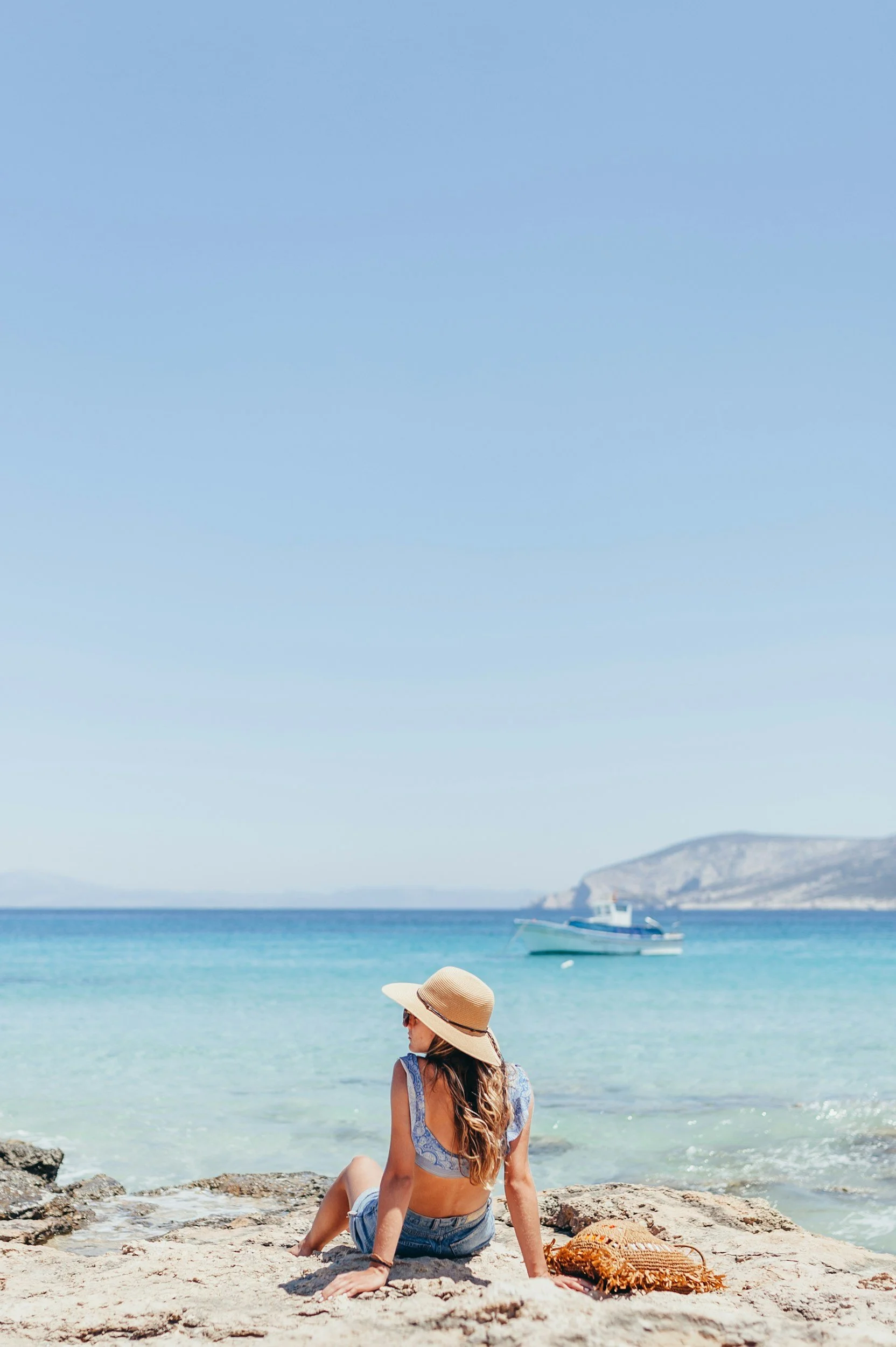 A woman sitting on a rocky beach, wearing a sun hat and sunglasses, looking at the ocean with a boat in the water and mountains in the background.