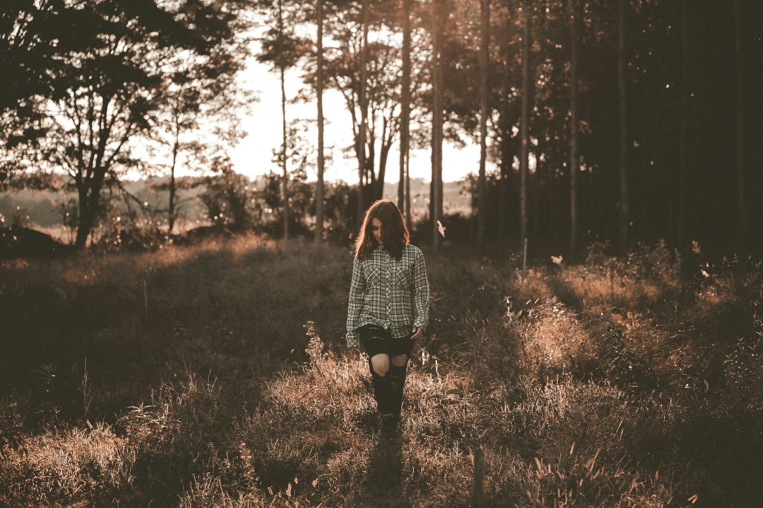 Young woman with dark hair, reflecting on her childhood wounds, walking through a field of tall grass and wildflowers during sunset, in the woods. The girl appears to be calm and grounded after Trauma therapy in Owings Mills, MD