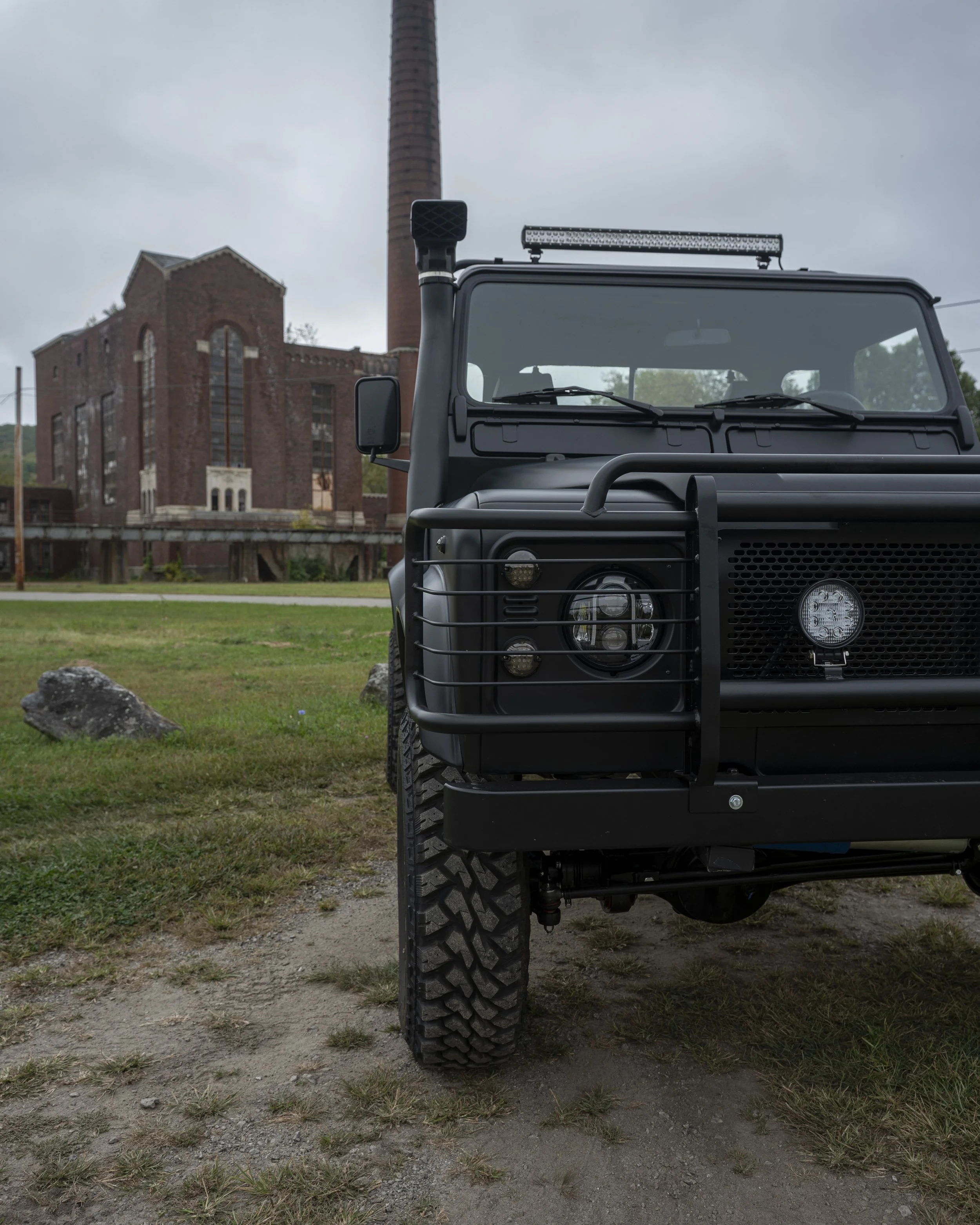 Front view of a black off-road vehicle with a protective grille on the front, parked on grass and dirt in front of an old brick factory with tall chimney in cloudy weather.