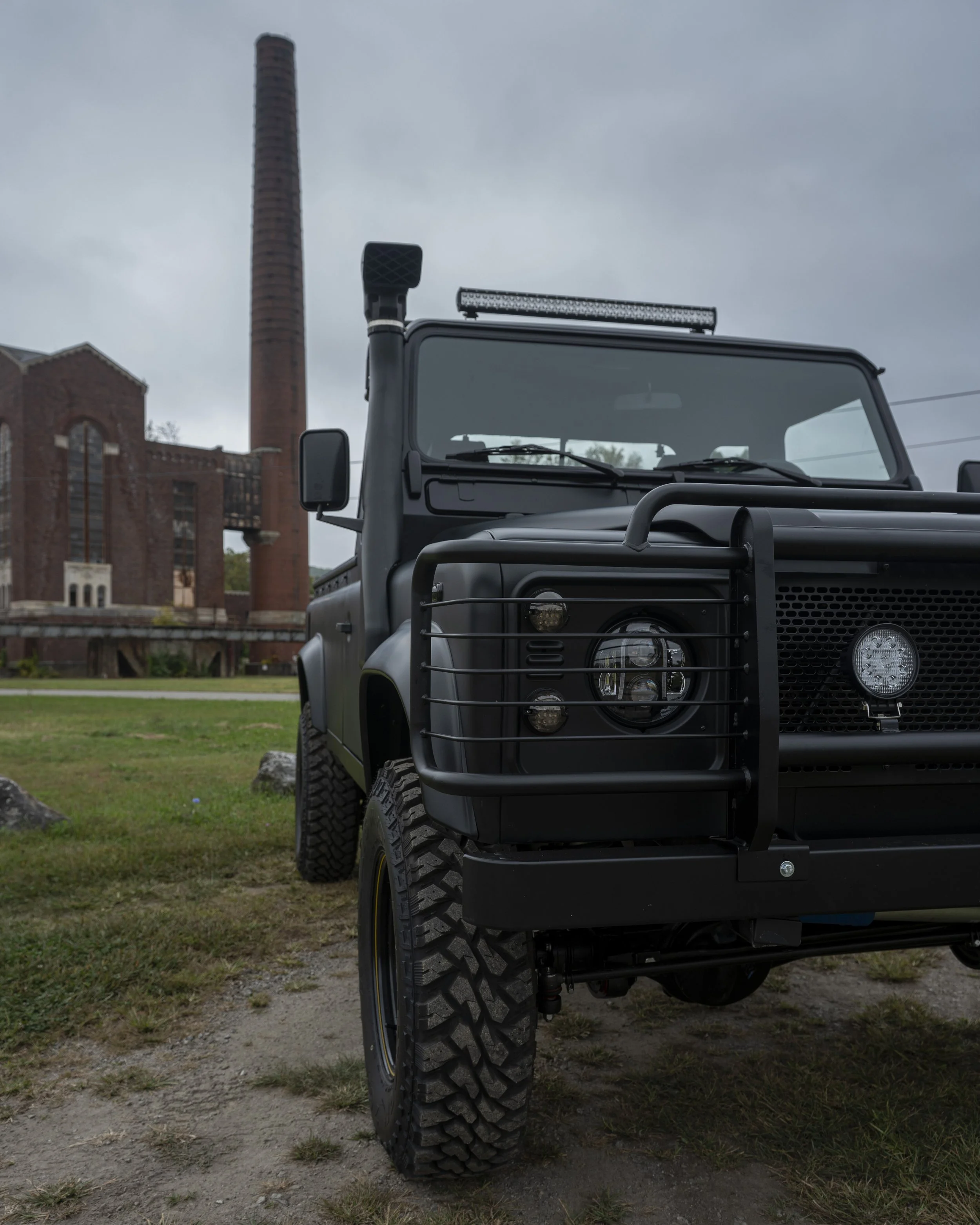 A black off-road vehicle with a large front grill guard, rugged tires, and a roof-mounted light bar, parked on a grassy area with an old industrial building and a tall smokestack in the background under a cloudy sky.