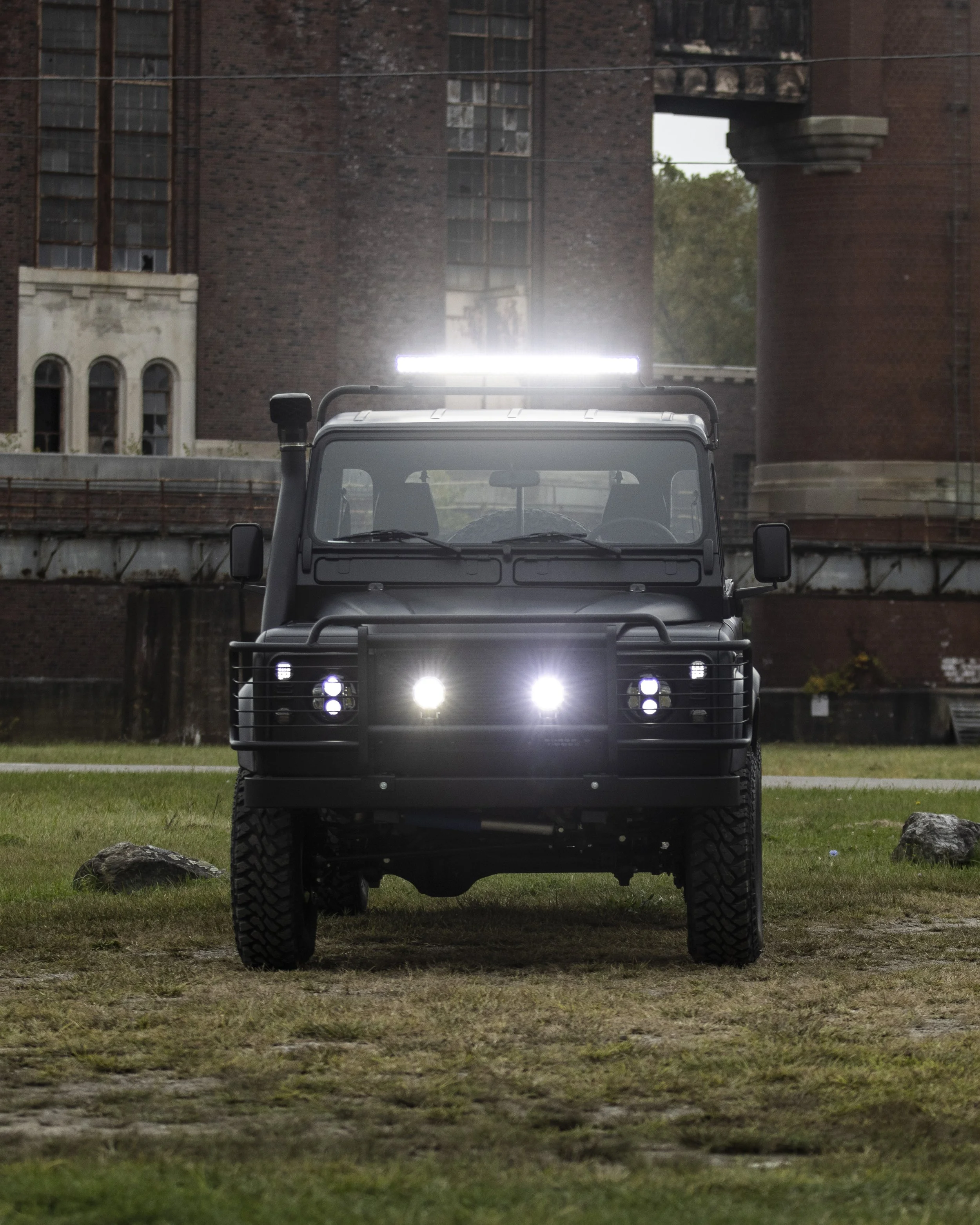 Black off-road vehicle with front grille guard and multiple headlights, parked on grass with old brick building in background.