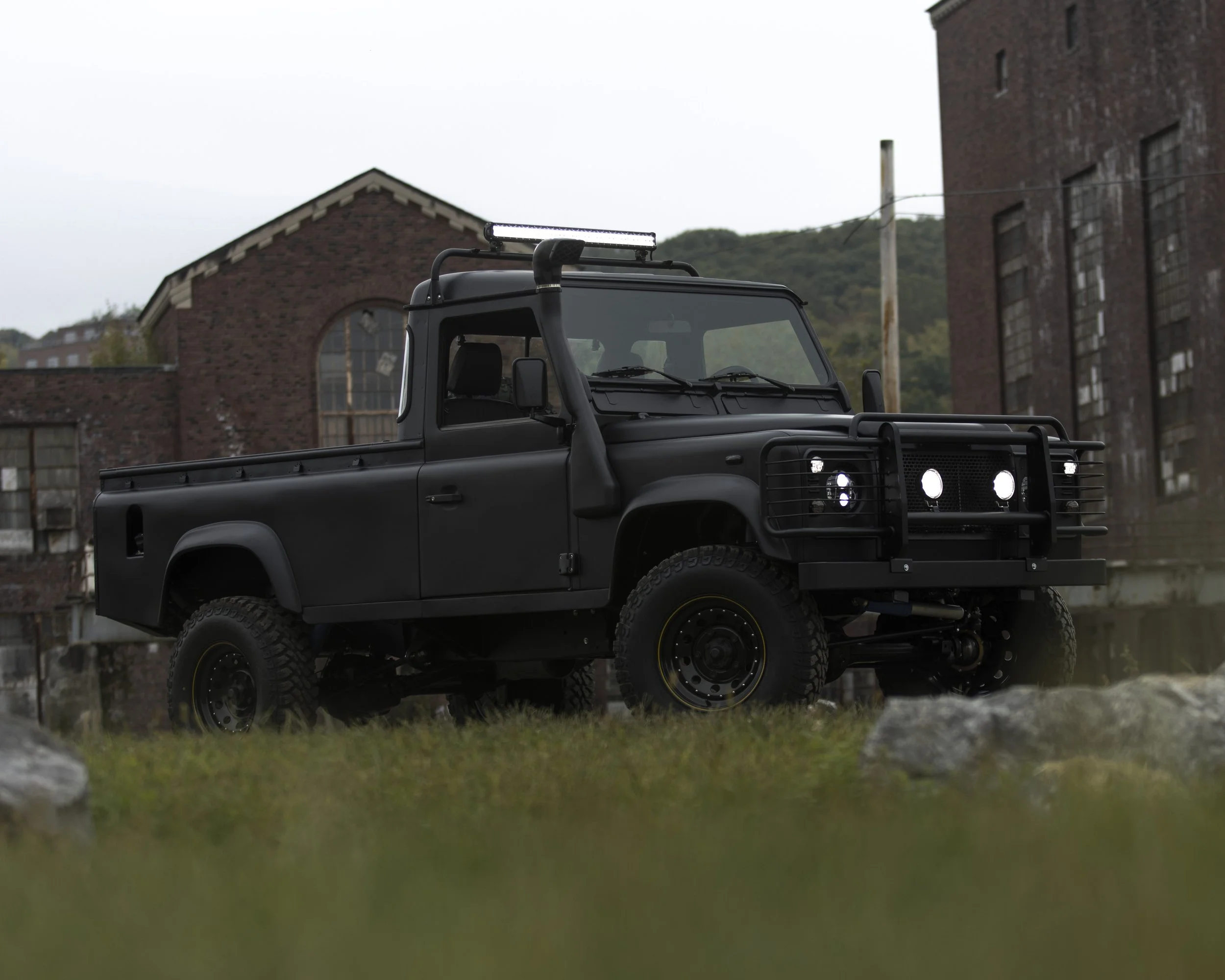 A black off-road pickup truck parked on a grassy area near an industrial building, with a mountain in the background.
