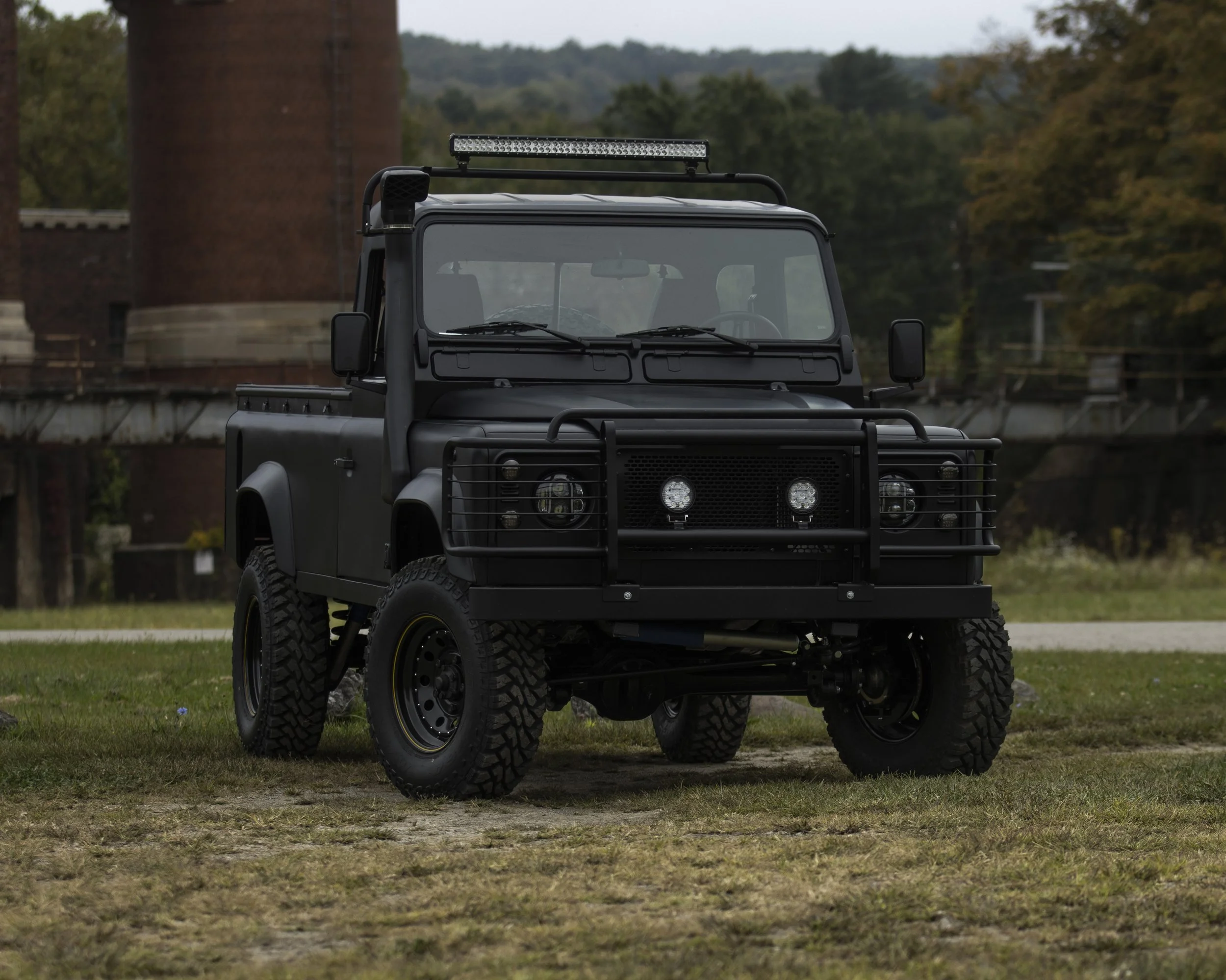 A black, rugged off-road utility vehicle with large tires and protective front grill, parked on grass with industrial structures and trees in the background.