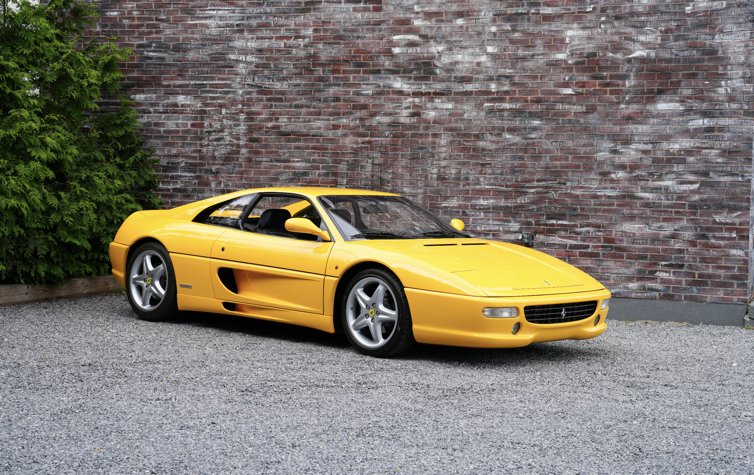 A yellow Ferrari sports car parked on a gravel driveway next to a brick wall and green bushes.