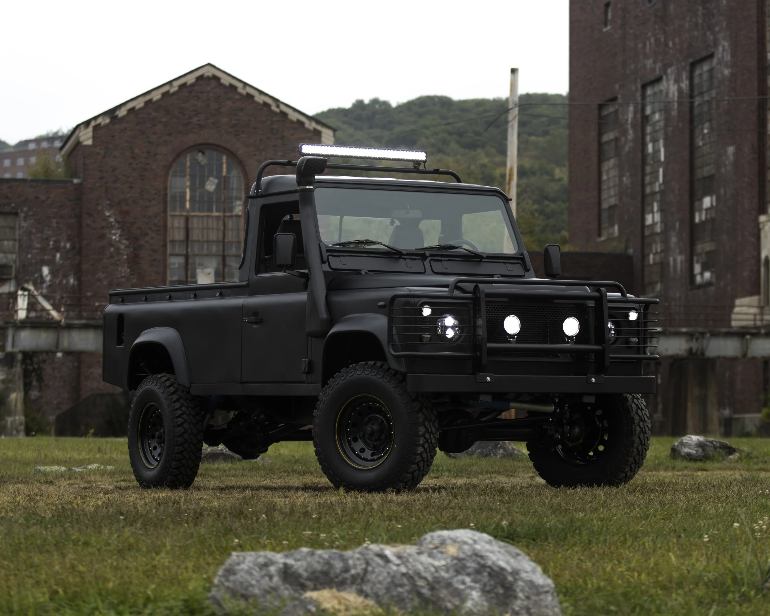 Black off-road truck with a front grille guard, roof light bar, and rugged tires parked on grass with factory buildings and a hill in the background.