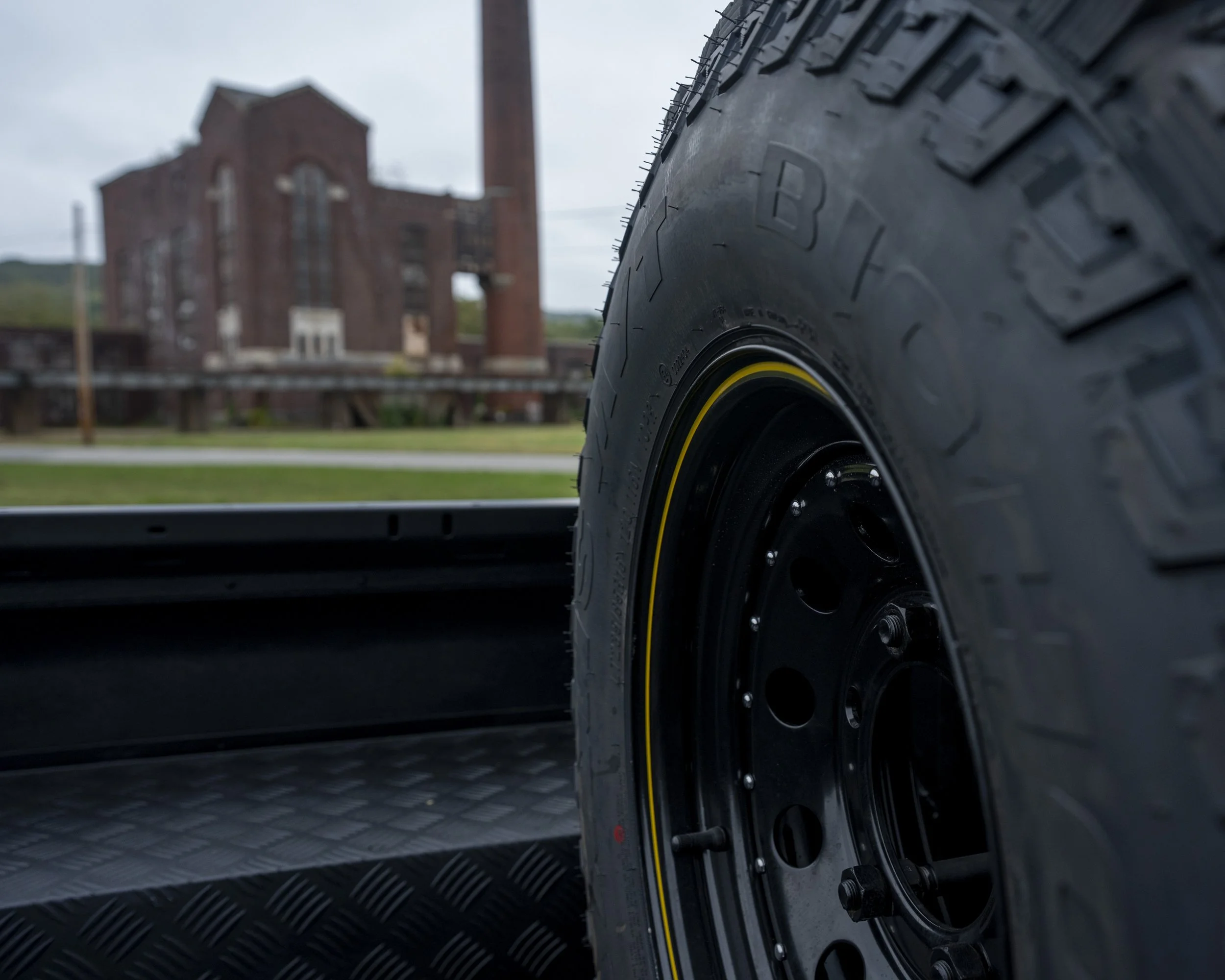 Close-up of a black tire with a yellow stripe on a matte black vehicle bed, with an industrial brick building in the background under a cloudy sky.