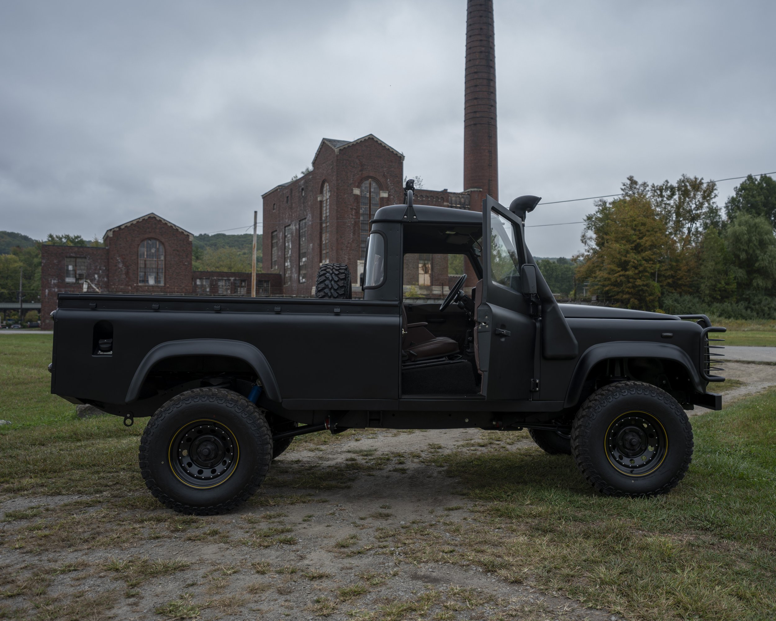 Black vintage off-road pickup truck parked on grass with an old industrial brick building and chimney in the background under cloudy sky.