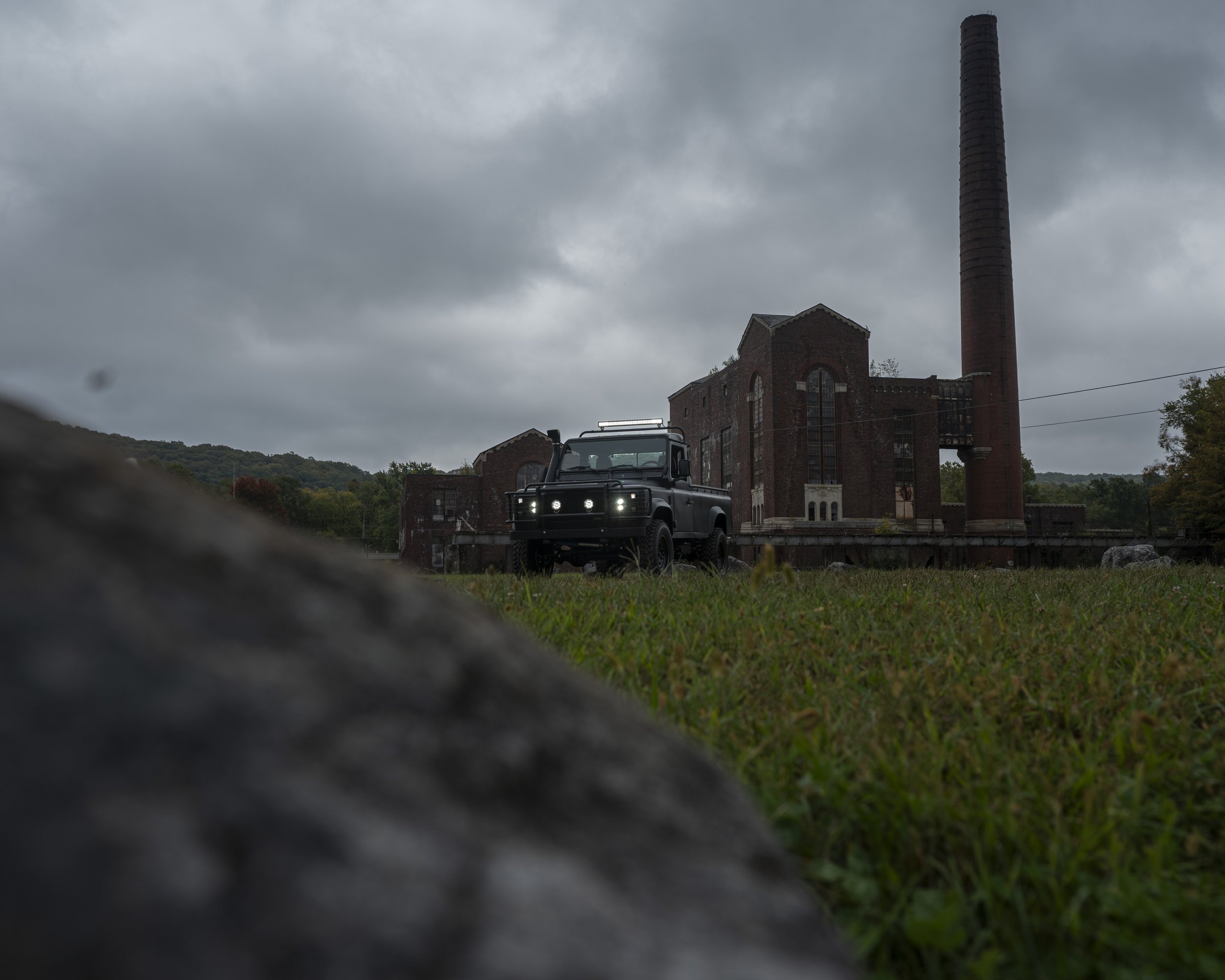A black off-road vehicle parked in front of an old red brick factory with a tall chimney, under cloudy skies, with grass in the foreground and hills in the background.