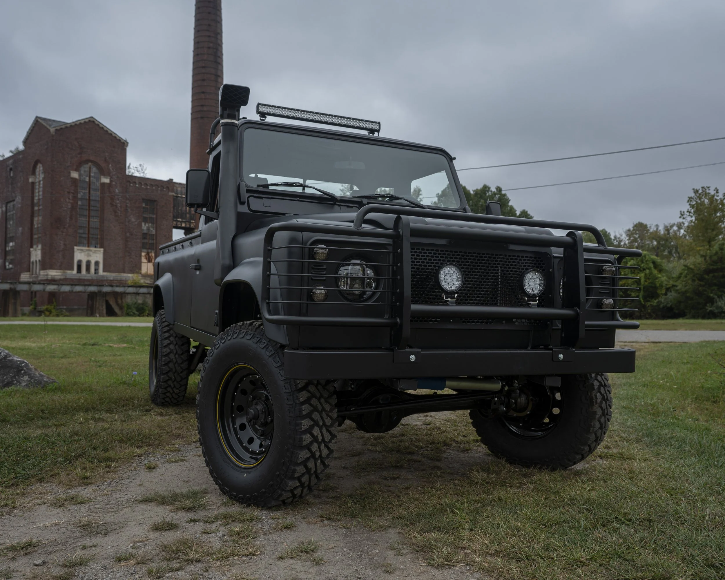 Black off-road vehicle with protective grille on front, large tires, and mounted lights, parked on grassy area with industrial building and trees in background under cloudy sky.