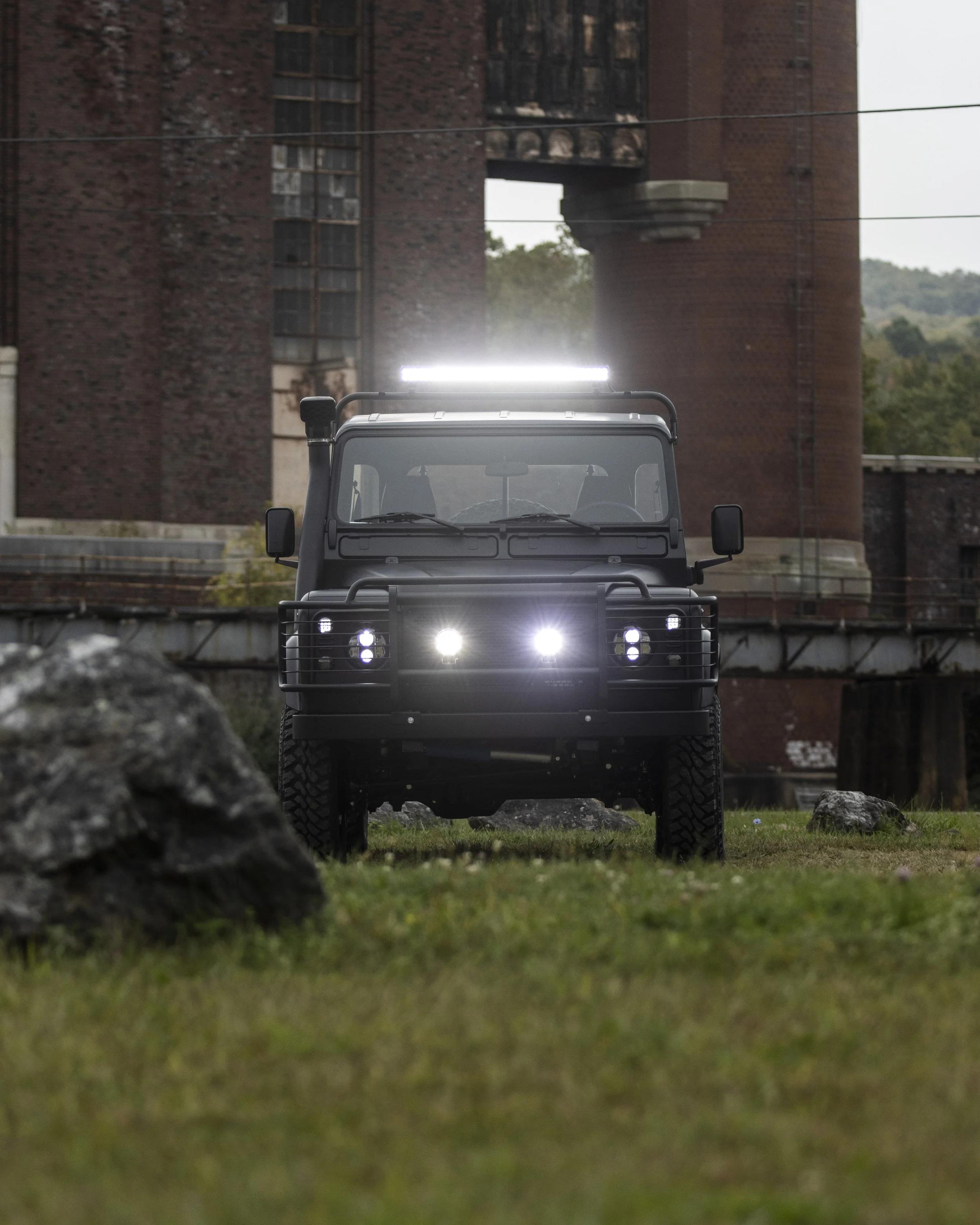 Black off-road vehicle with bright front and roof lights facing forward on grassy terrain in front of a large, old brick building.