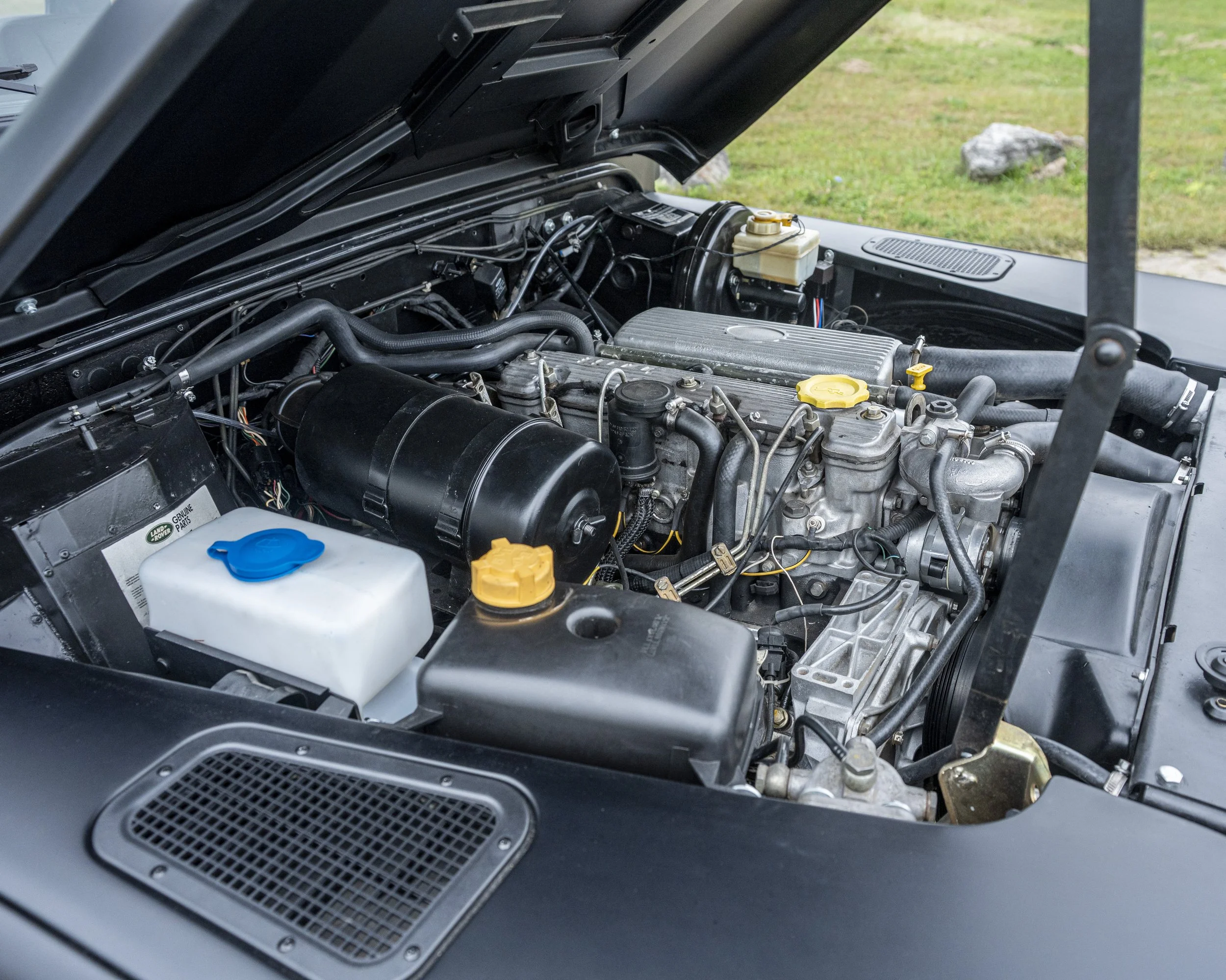 Close-up of a car engine compartment with various mechanical parts, hoses, and fluid reservoirs visible.