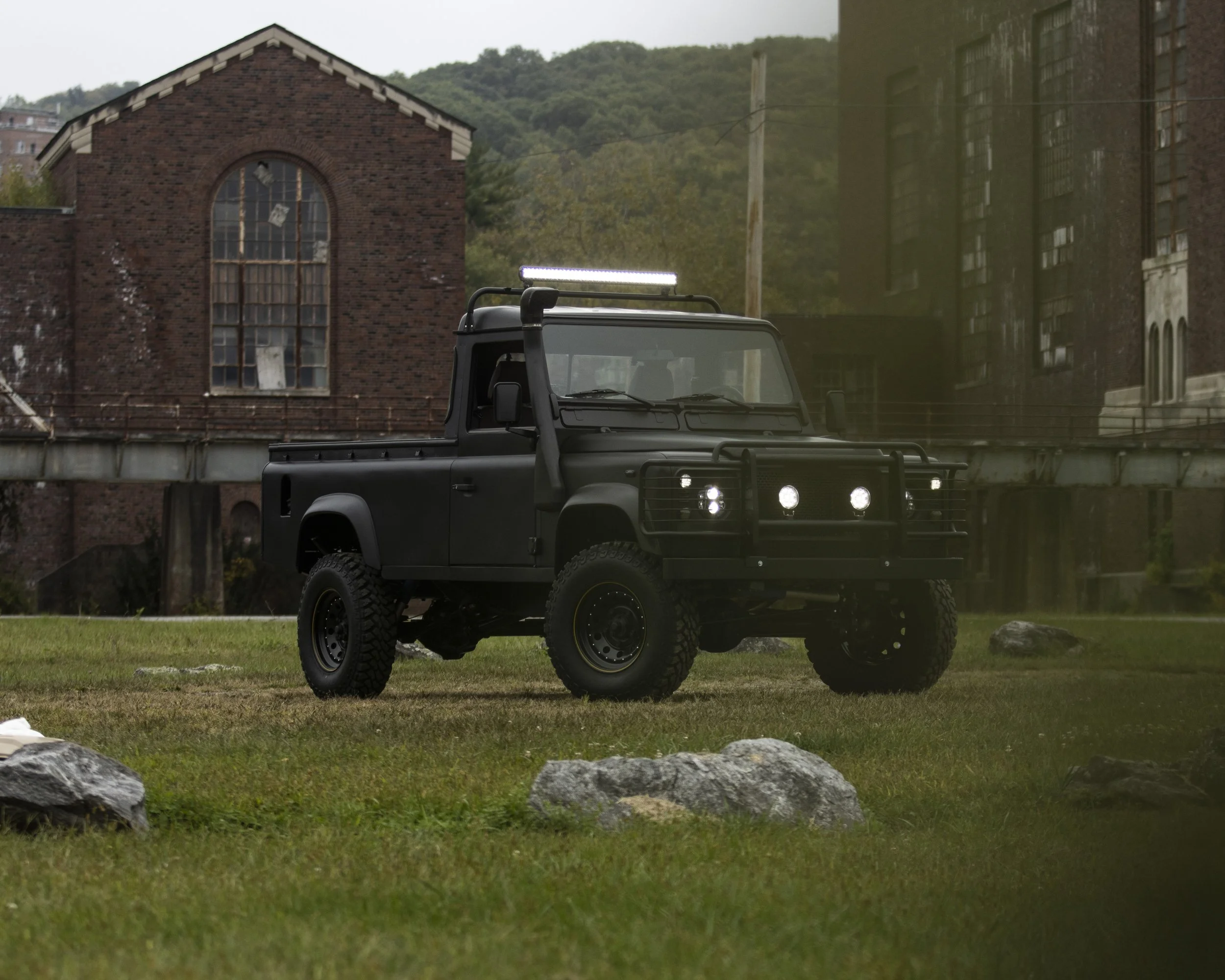 A black off-road utility vehicle with rugged tires, LED light bar, and front grill guard parked on a grassy area with rocks, in front of a background of old brick buildings and a hillside.