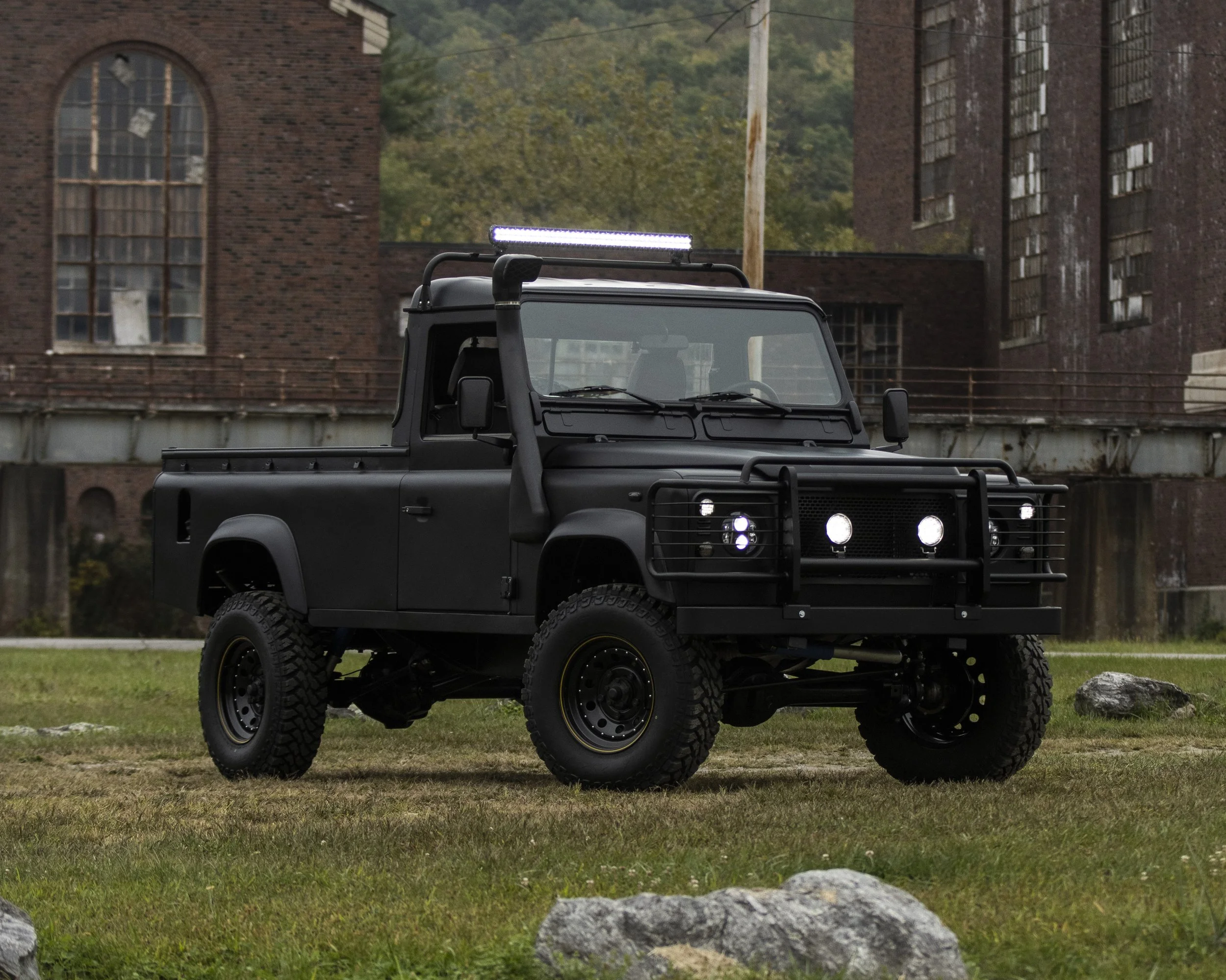 Black off-road utility vehicle with rugged tires, front grille guard, and roof-mounted light bar parked on grassy area with old brick buildings in the background.