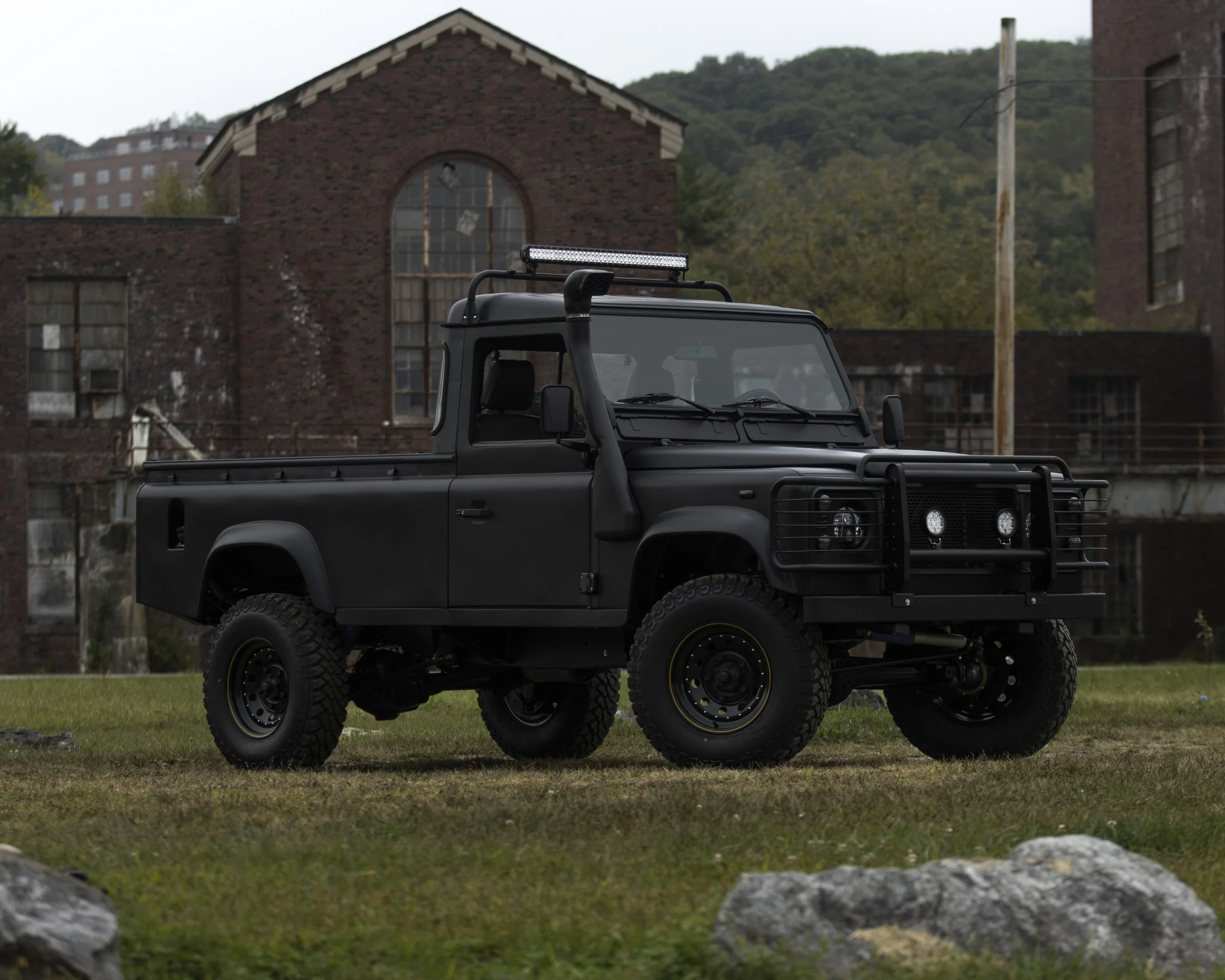 Black off-road utility vehicle with LED light bar, parked on grassy area in front of old brick industrial buildings.