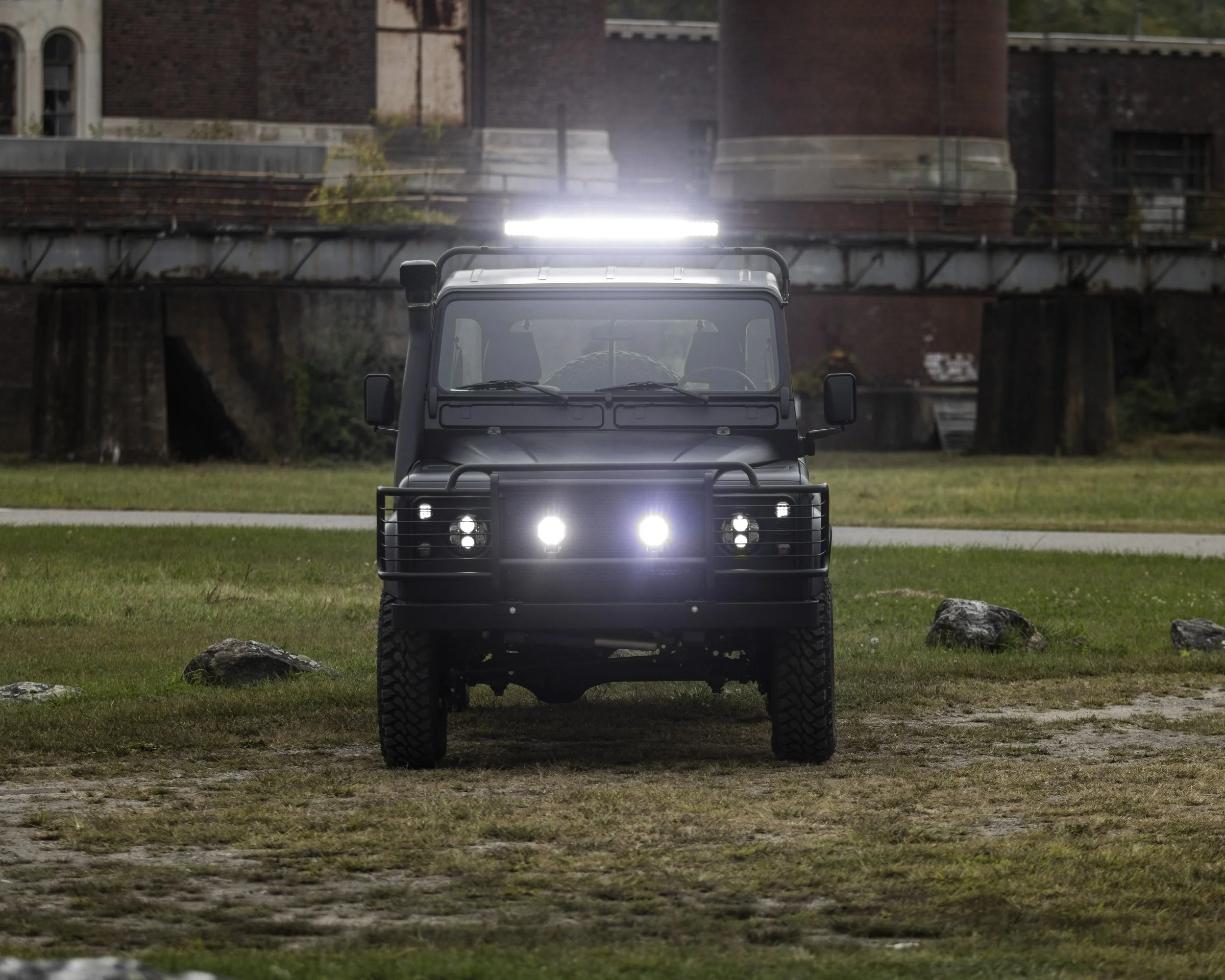 A black off-road vehicle with bright headlights facing forward on a grassy area with rocks, background includes a bridge and brick buildings.