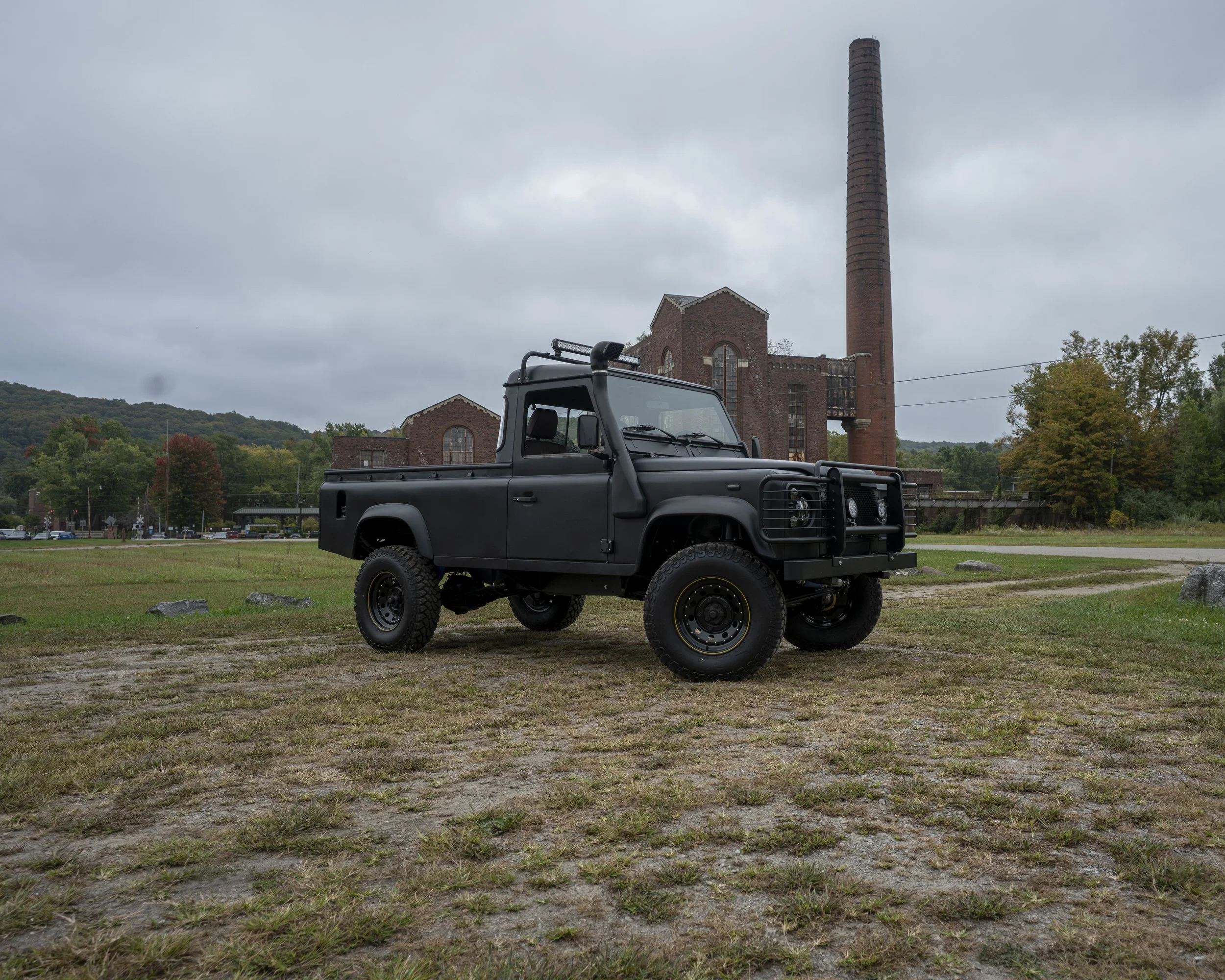 A black off-road vehicle parked on a grassy field with a historic brick factory building and large smokestack in the background on a cloudy day.