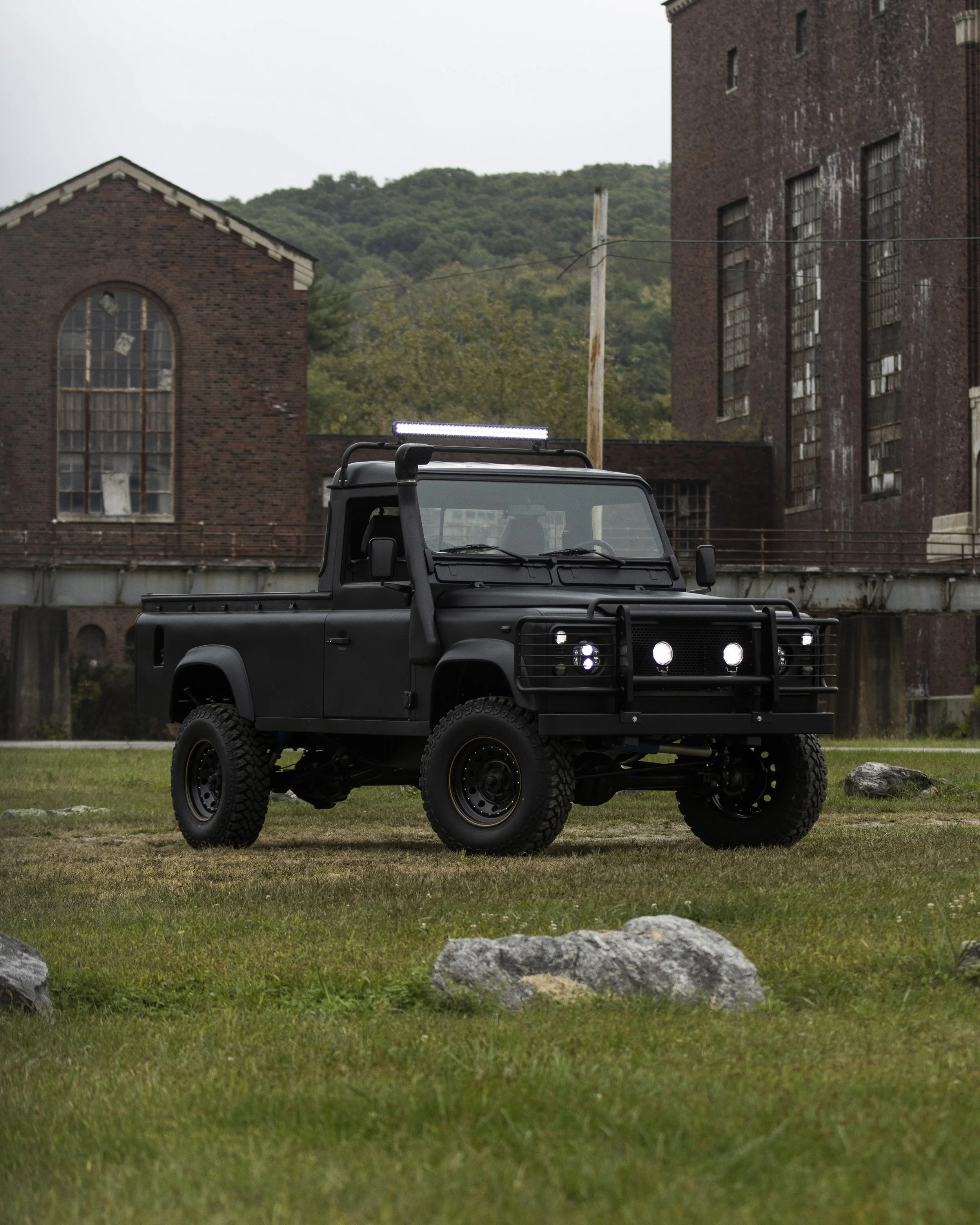 Black off-road truck parked on grass with abandoned brick buildings and a mountain in the background