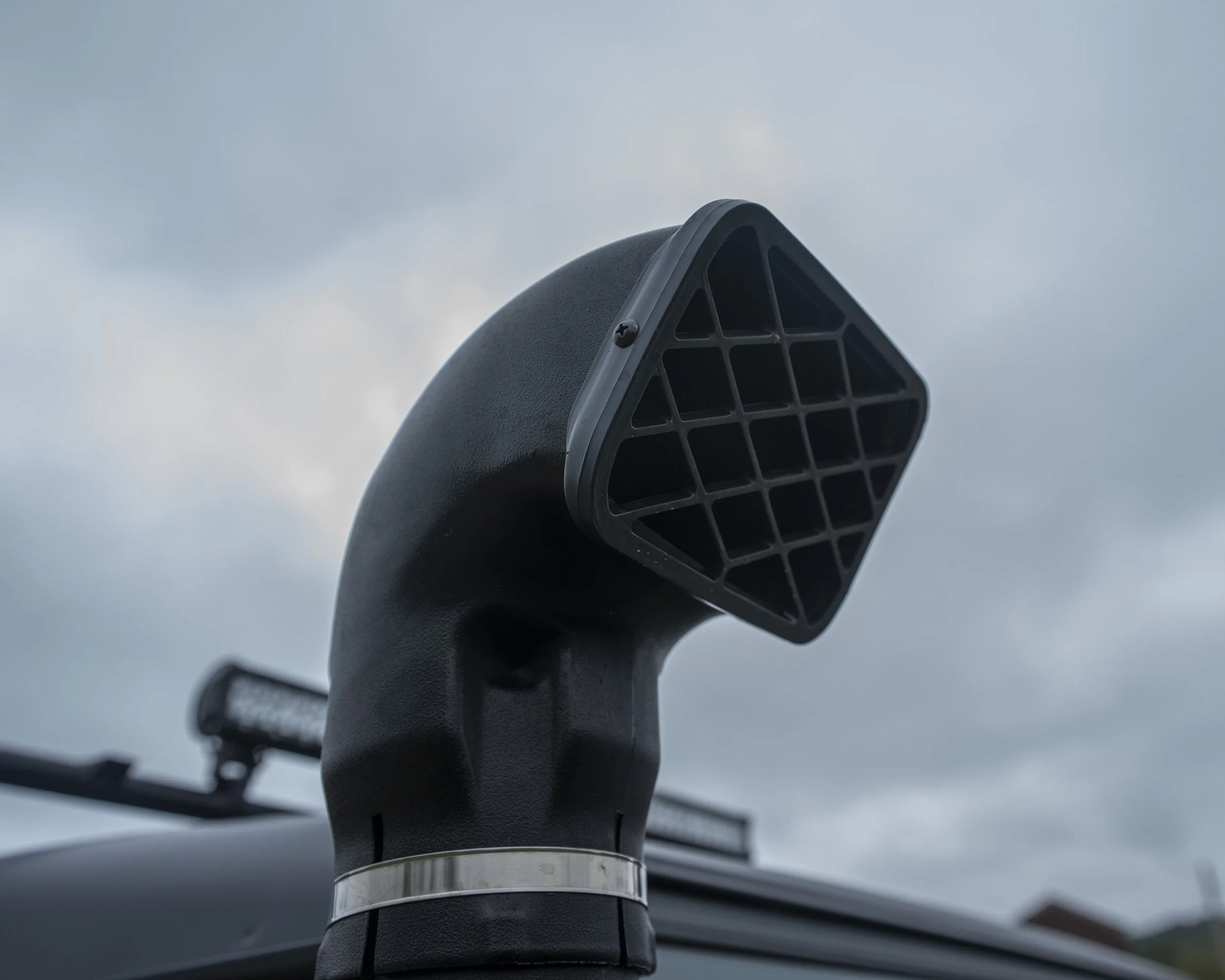 Close-up of a black vehicle snorkel with a square air intake, against a cloudy sky.