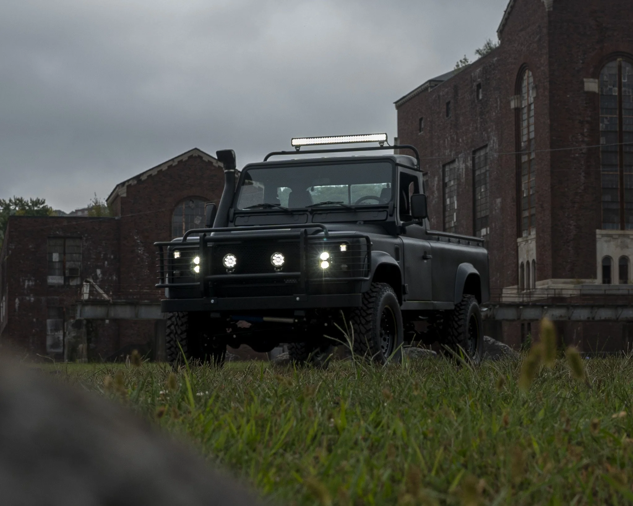 A black off-road vehicle with LED lights on the roof and front grille parked on a grassy field with a brick building in the background on a cloudy day.