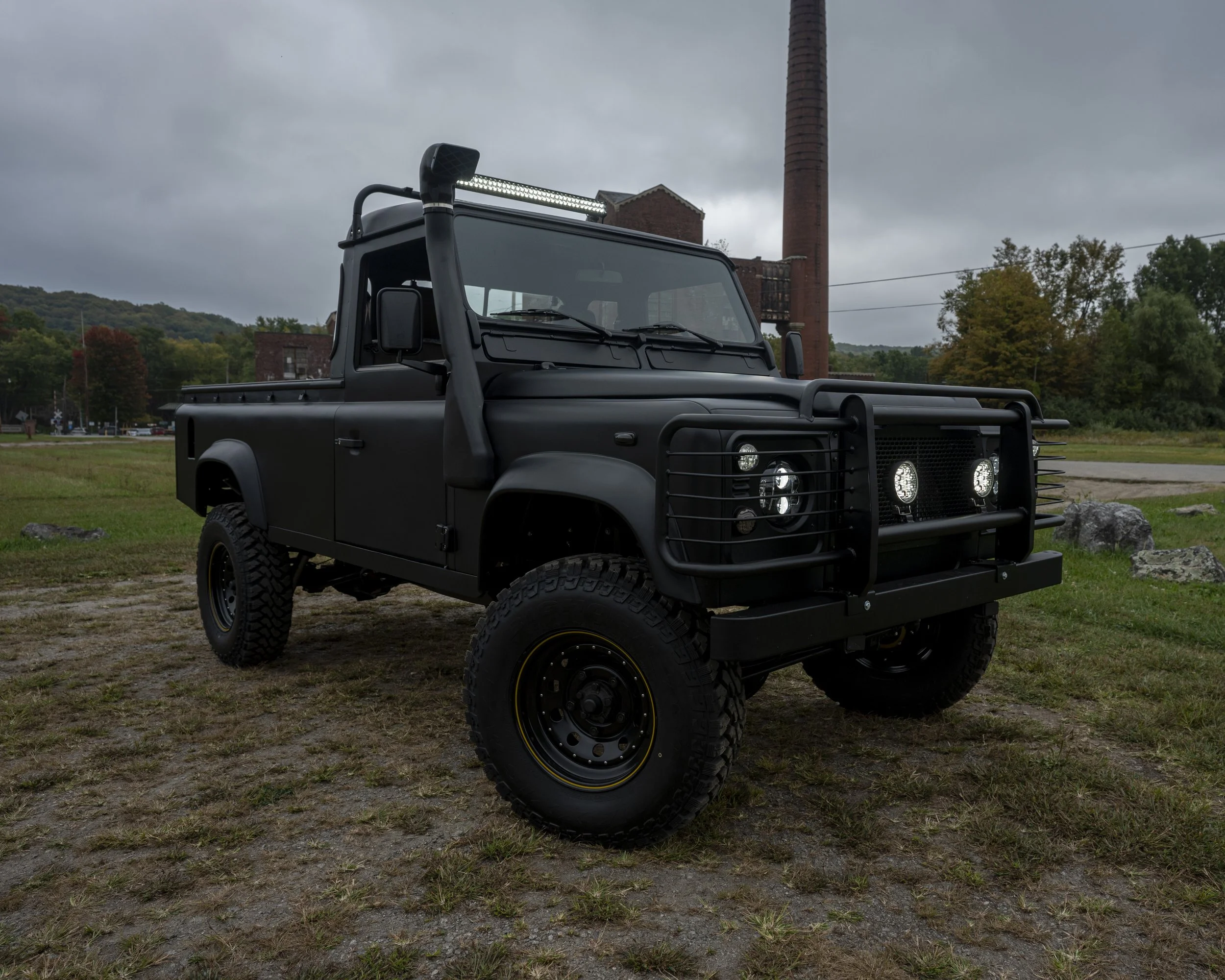 A black off-road utility vehicle with large tires, bumper guard, and LED light bar parked on grass with trees and an industrial building in the background.