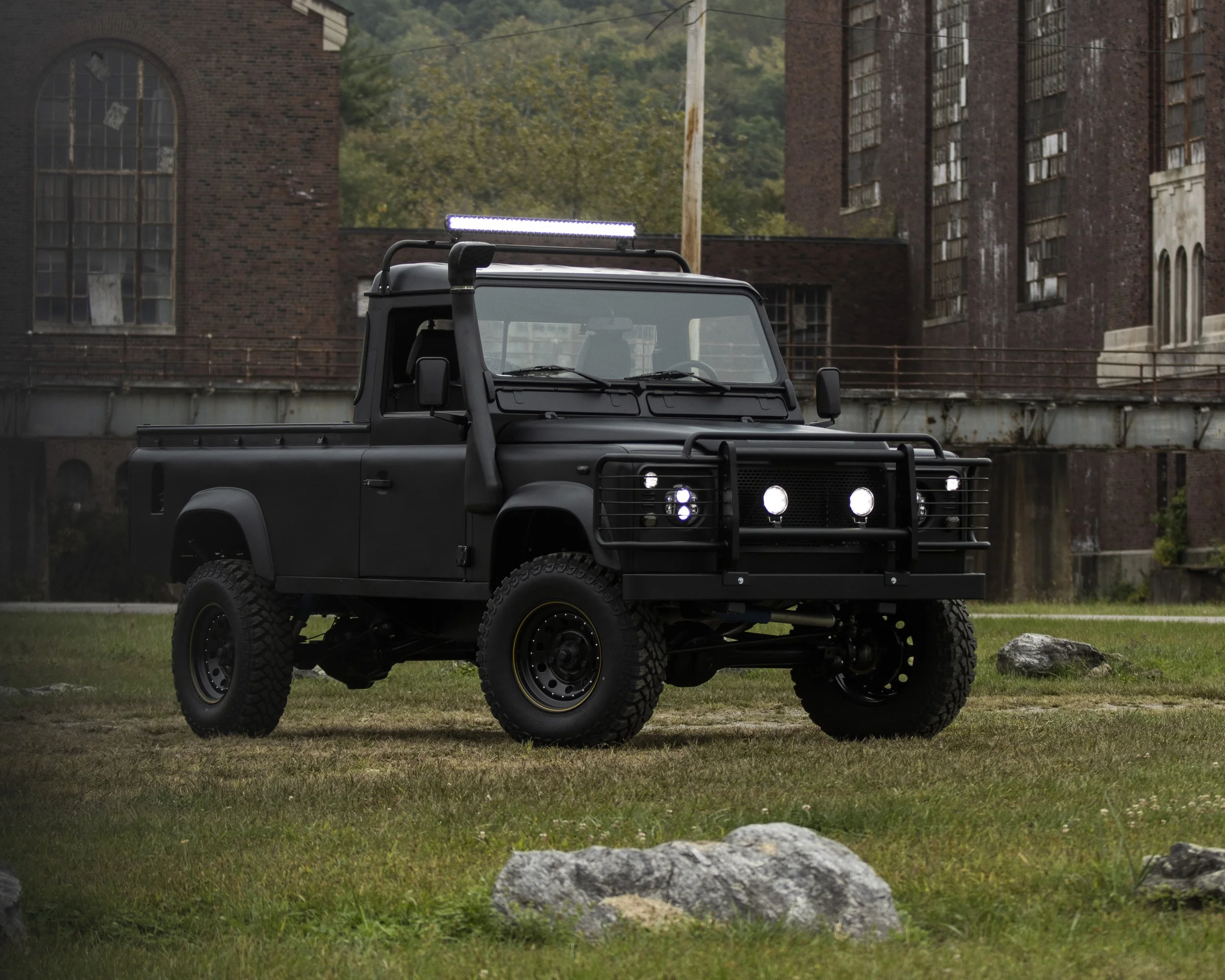 A black off-road utility vehicle with a matte finish parked on grass with rocks in front, behind an old industrial building with brick walls and large windows, and trees in the background.