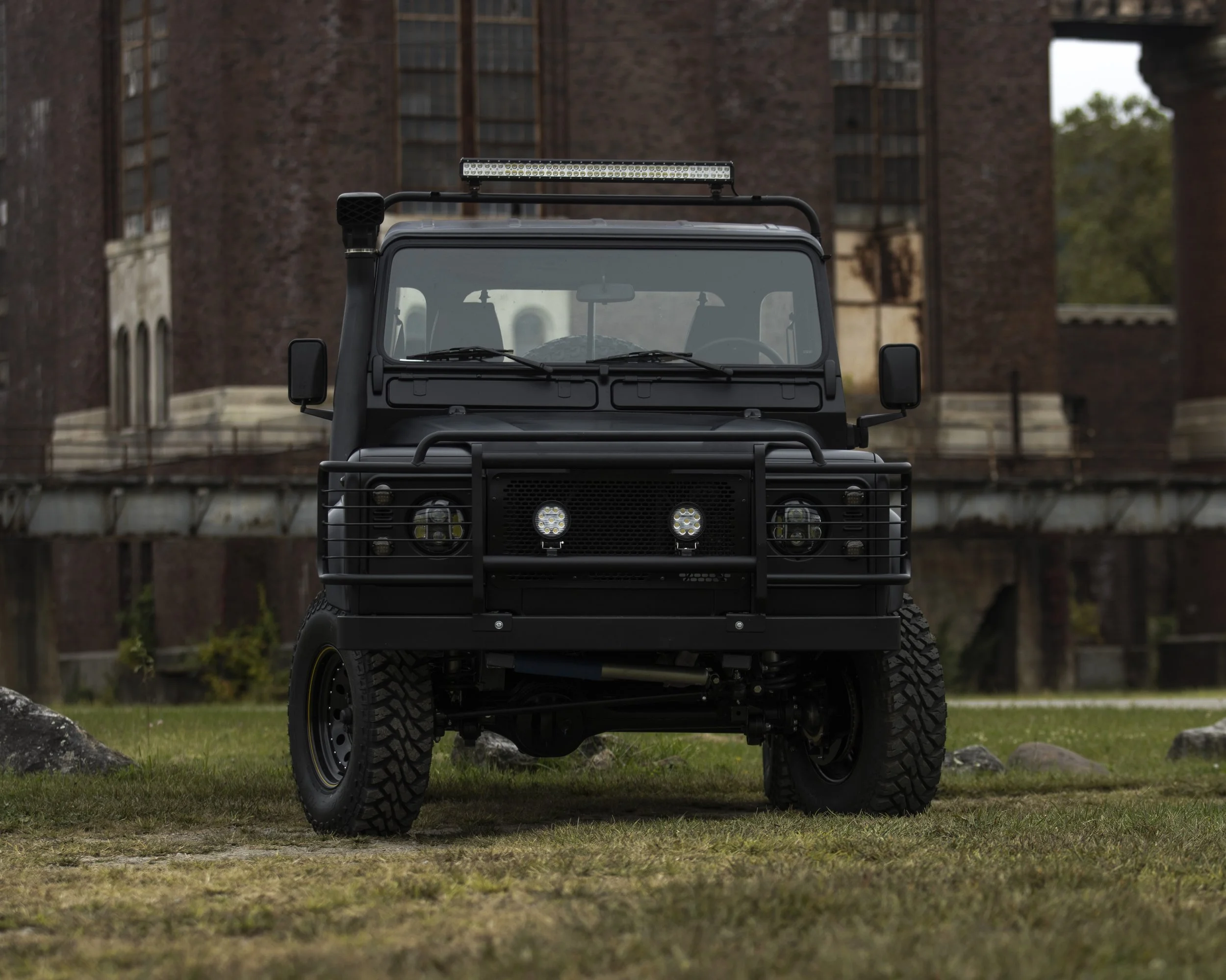 Black off-road vehicle with a metal roof rack and front grille guard, parked on grassy area with historic brick buildings in the background.