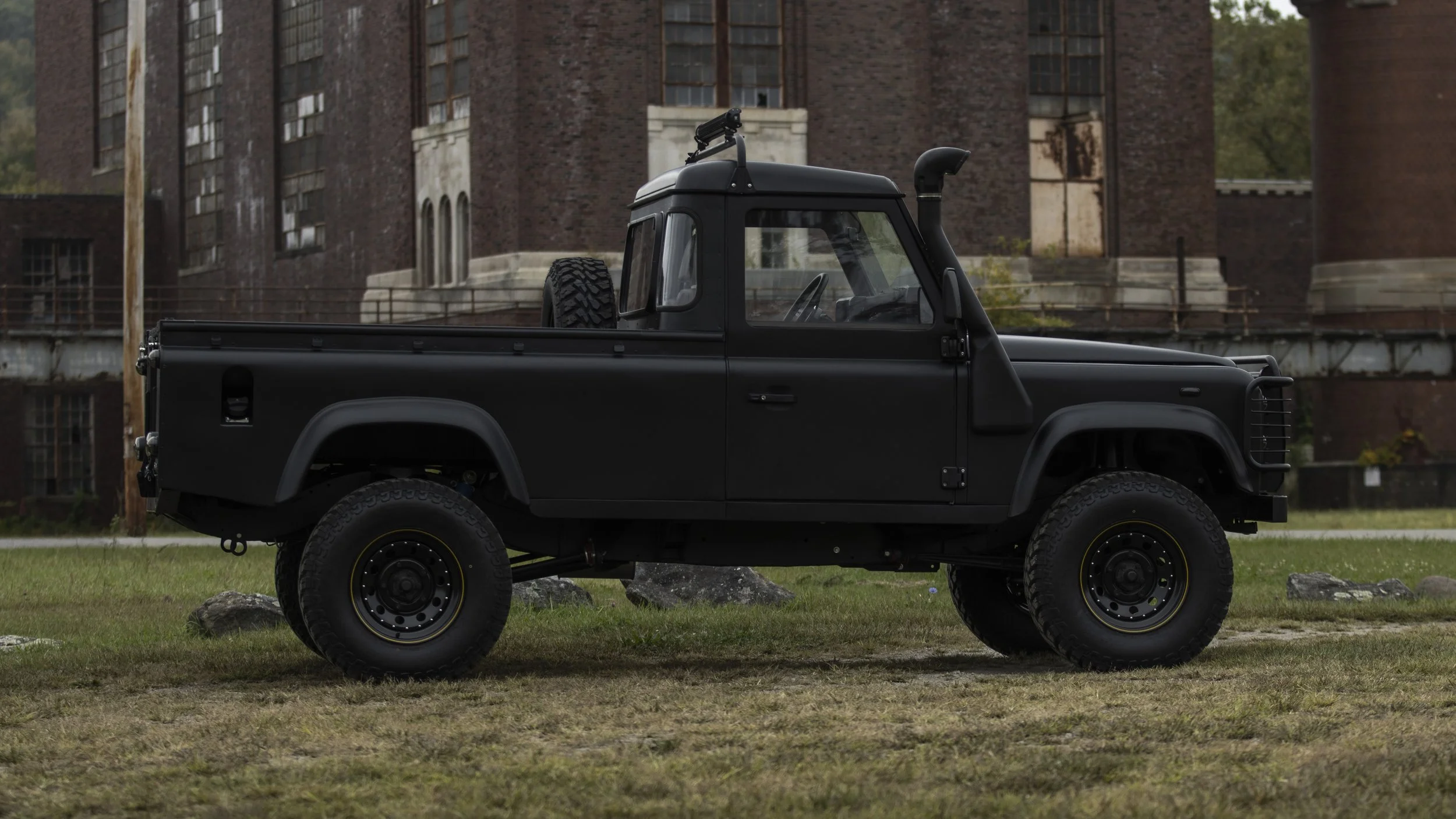 A black vintage pickup truck parked on a grassy field in front of an old brick building with large windows.