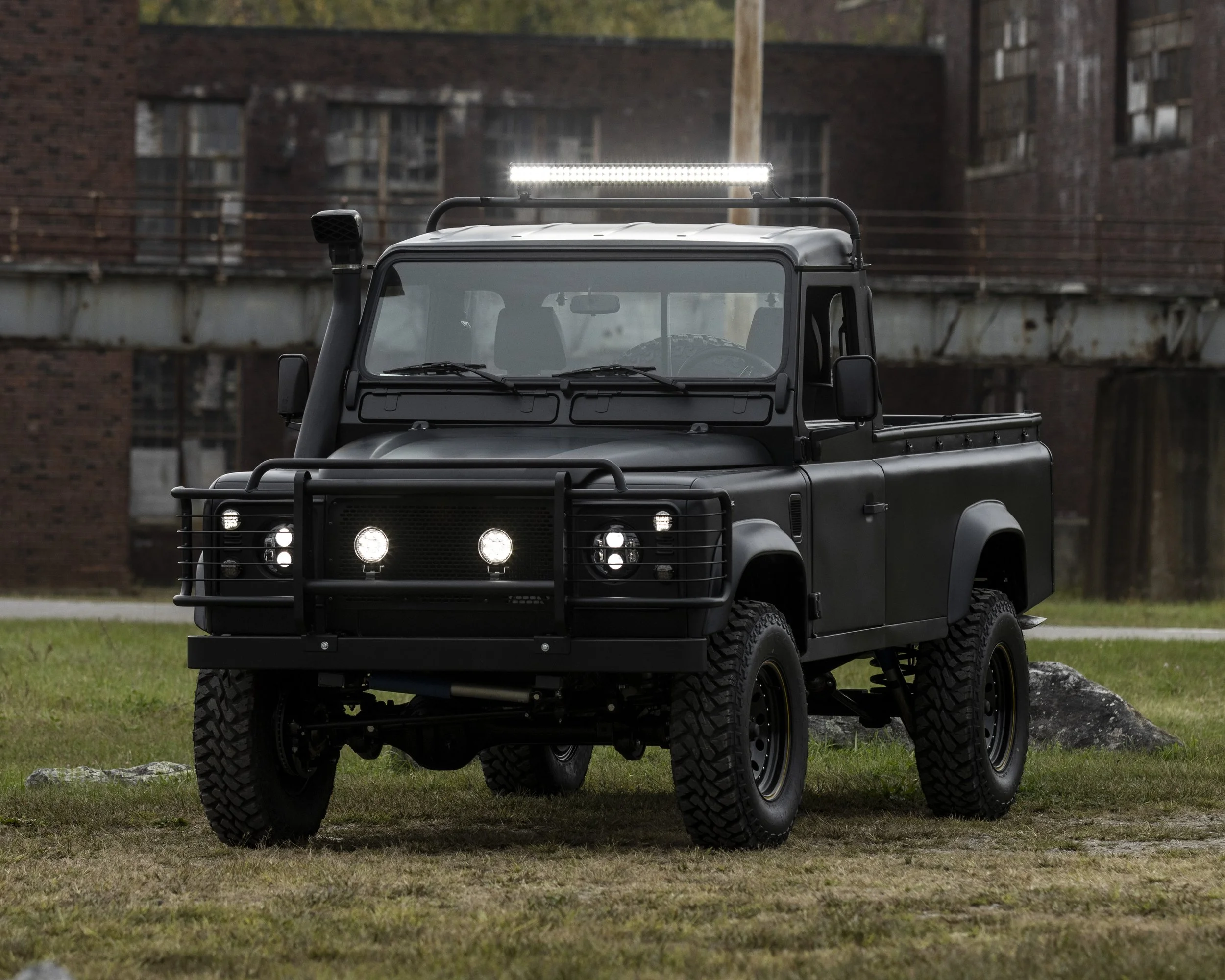 A black off-road utility vehicle with a roof light, front grille guard, and large tires parked on grass with an industrial building in the background.