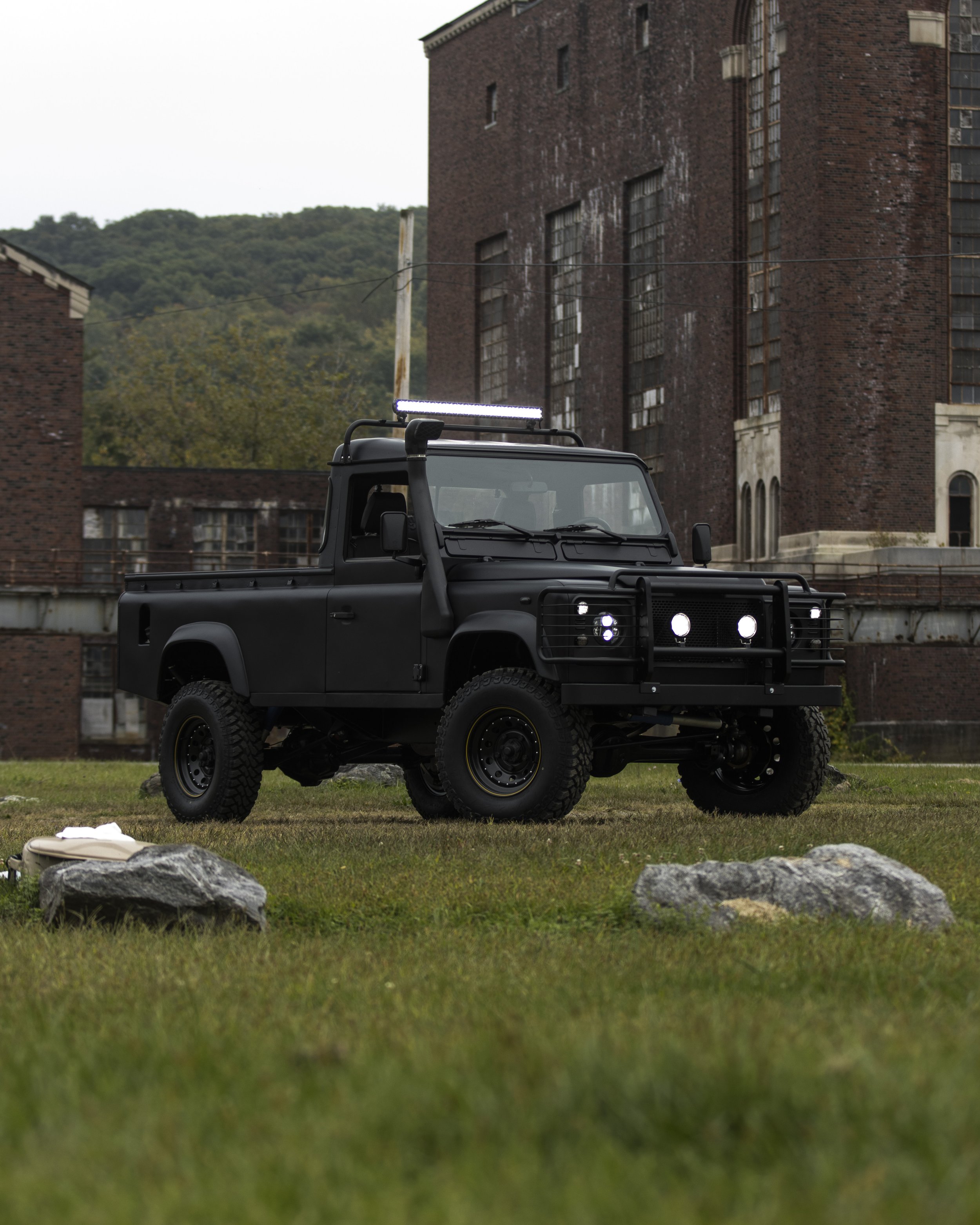 Black off-road vehicle with roof lights parked on grass near rocks in front of a brick building.