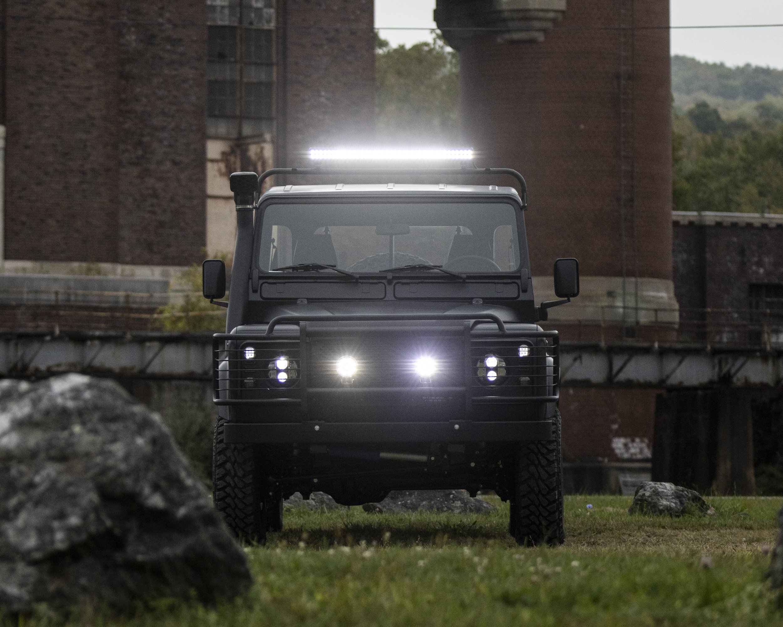 A black off-road utility vehicle with bright headlights and a rooftop light, positioned outdoors on a grassy area with rocks, in front of large industrial brick buildings and a bridge.
