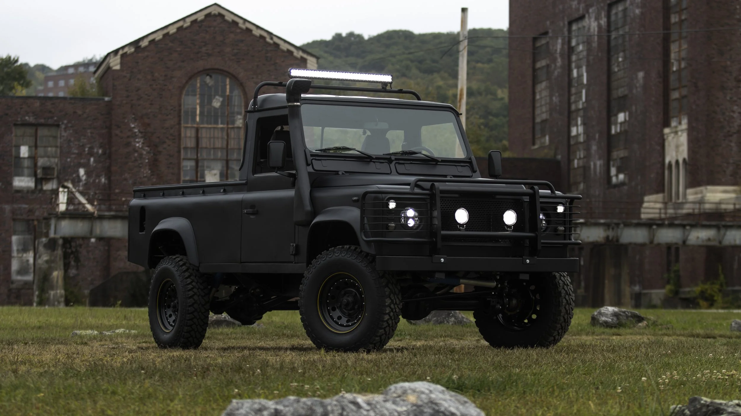 Black off-road vehicle with rugged tires and a protective front grill parked on grass in front of old red brick buildings.