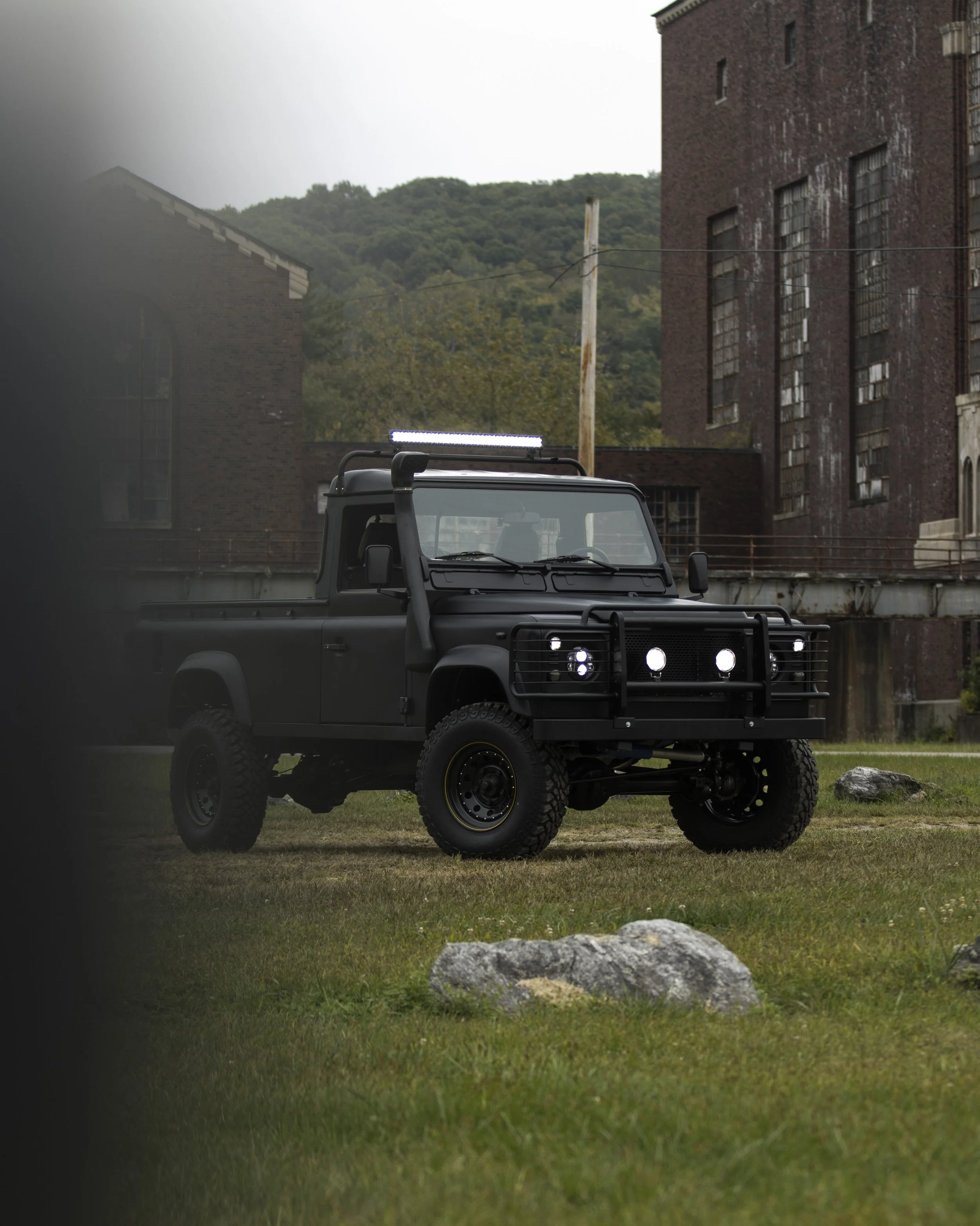 A black off-road vehicle, possibly a truck or SUV, parked on grass with a rugged appearance. The vehicle has large tires, a front grille guard, mounted lights on the roof and front, and a matte finish. It is situated in front of old brick buildings a
