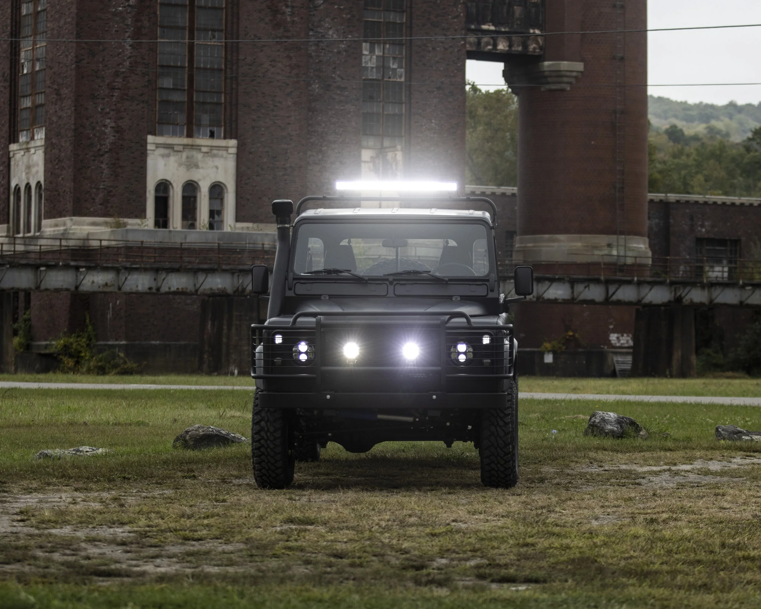 Black off-road utility vehicle with bright headlights, parked on grass in front of a historic brick building with towers and a bridge.
