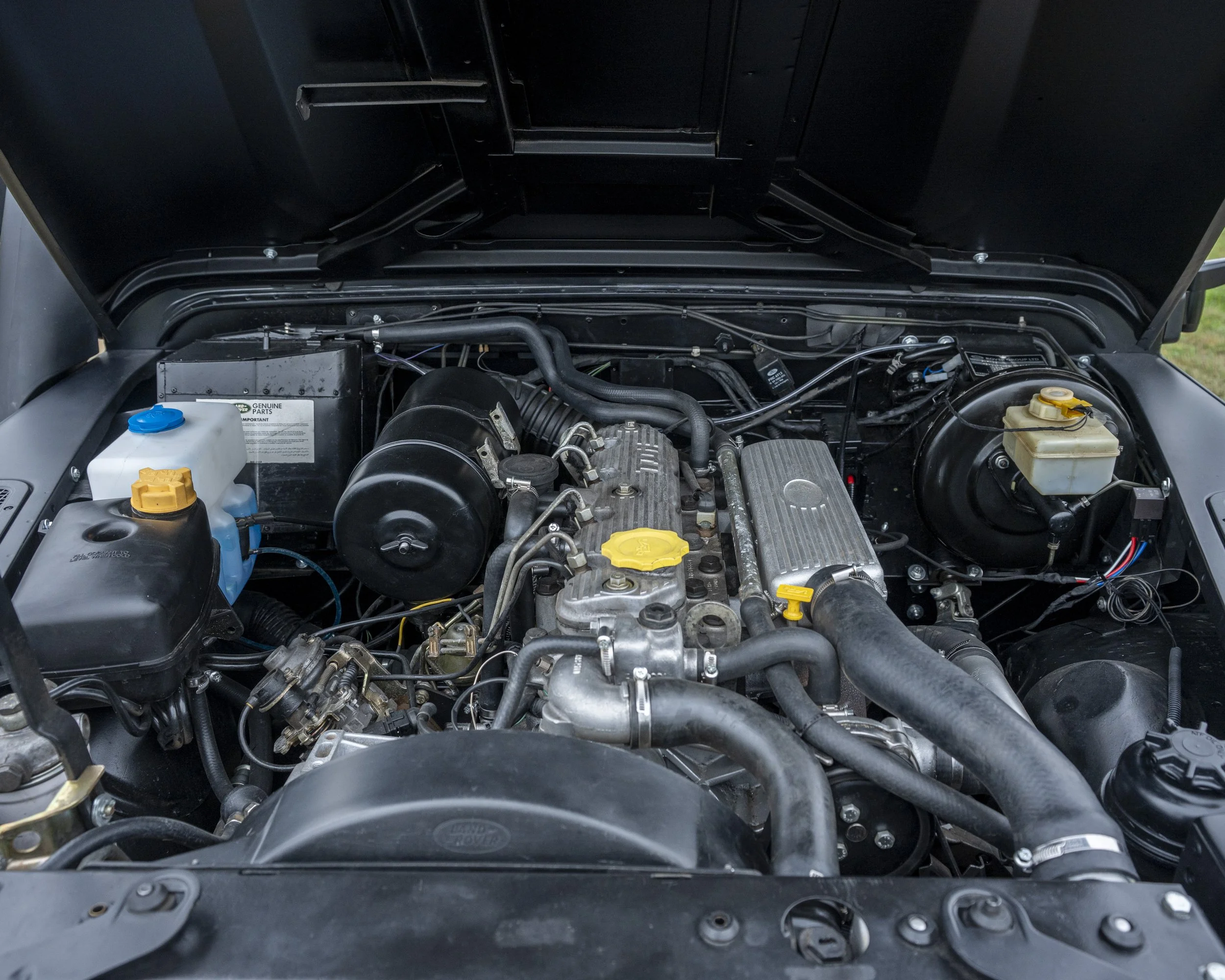 The image shows the engine bay of a vehicle with various components, including the engine, hoses, and fluid reservoirs.