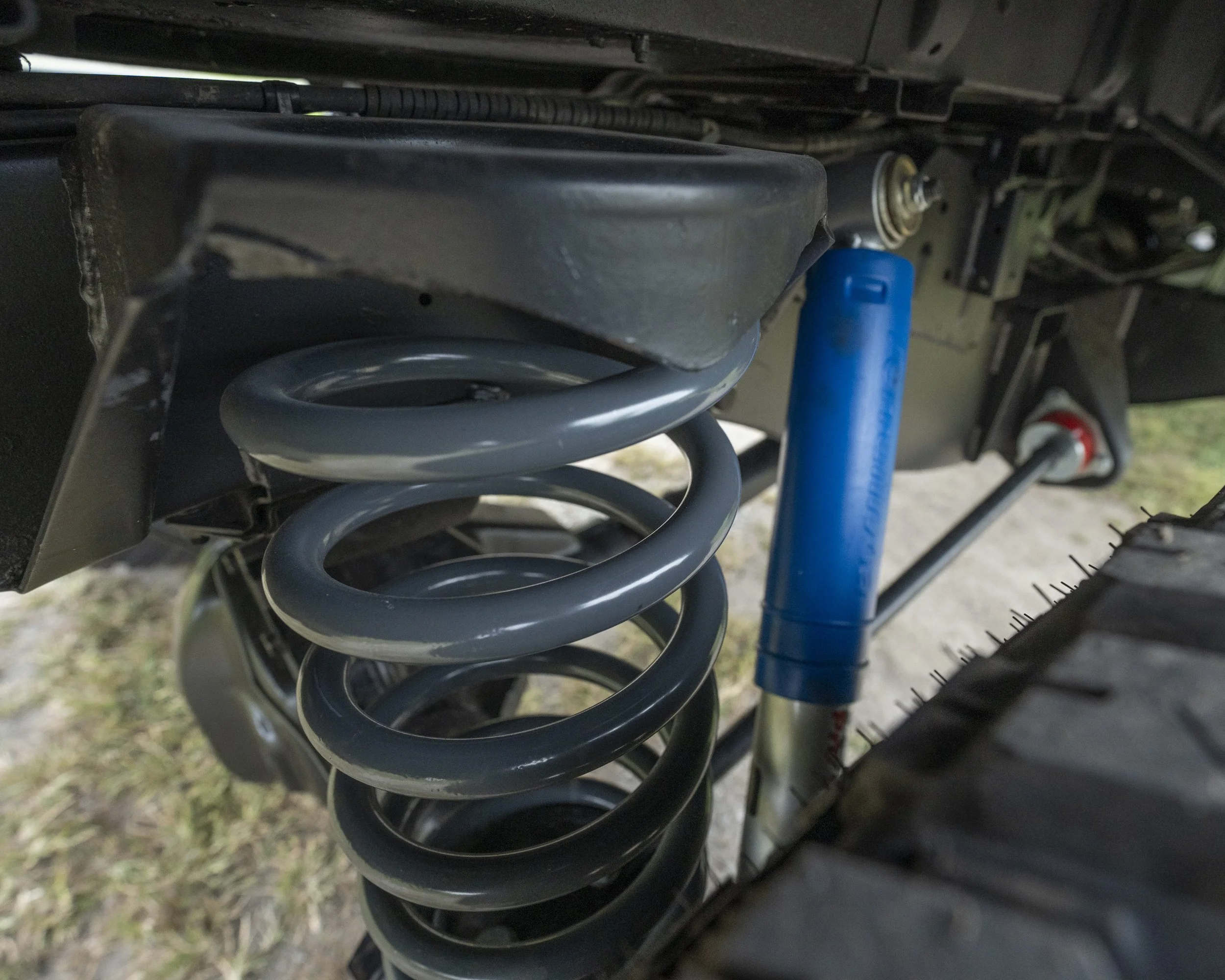 Close-up of vehicle suspension system showing a black coil spring, a blue shock absorber, and parts of the vehicle's undercarriage with a tire visible.