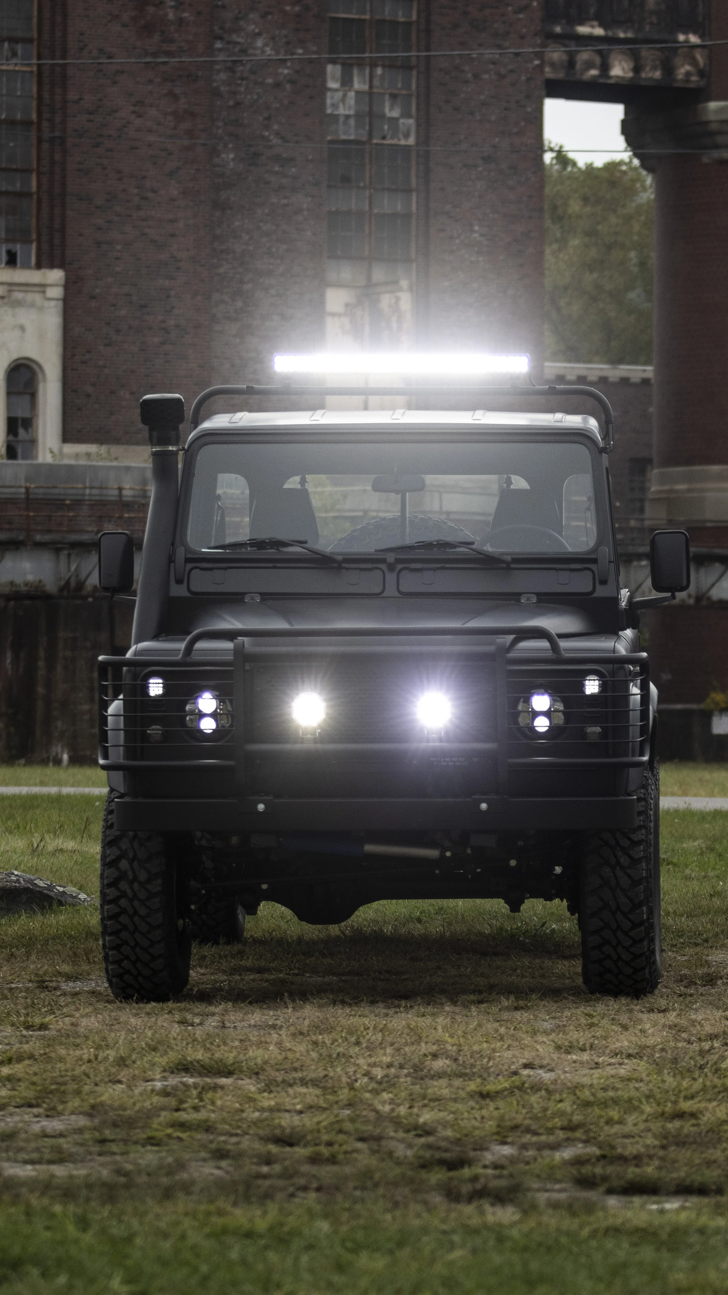 Front view of a black off-road vehicle with bright lights, parked on grass in an urban area with brick buildings in the background.