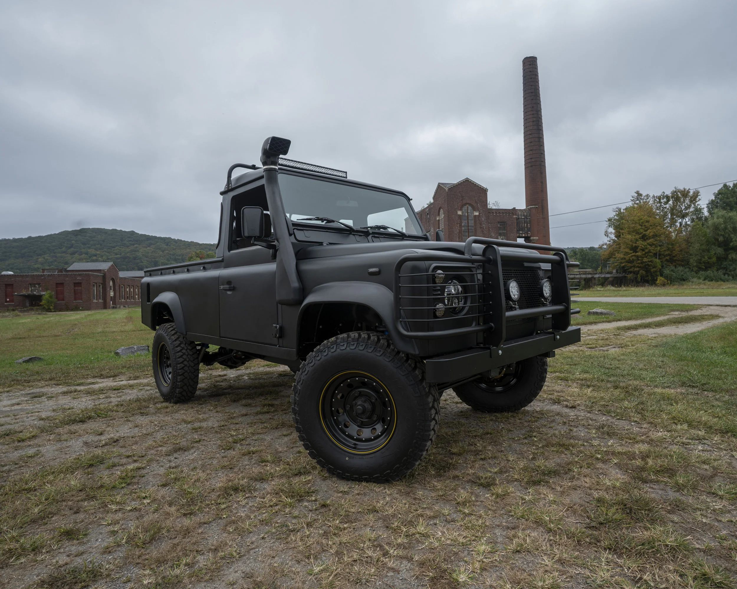 A black four-wheel-drive vehicle parked on a grassy area with old brick factory buildings and smokestack in the background under cloudy skies.