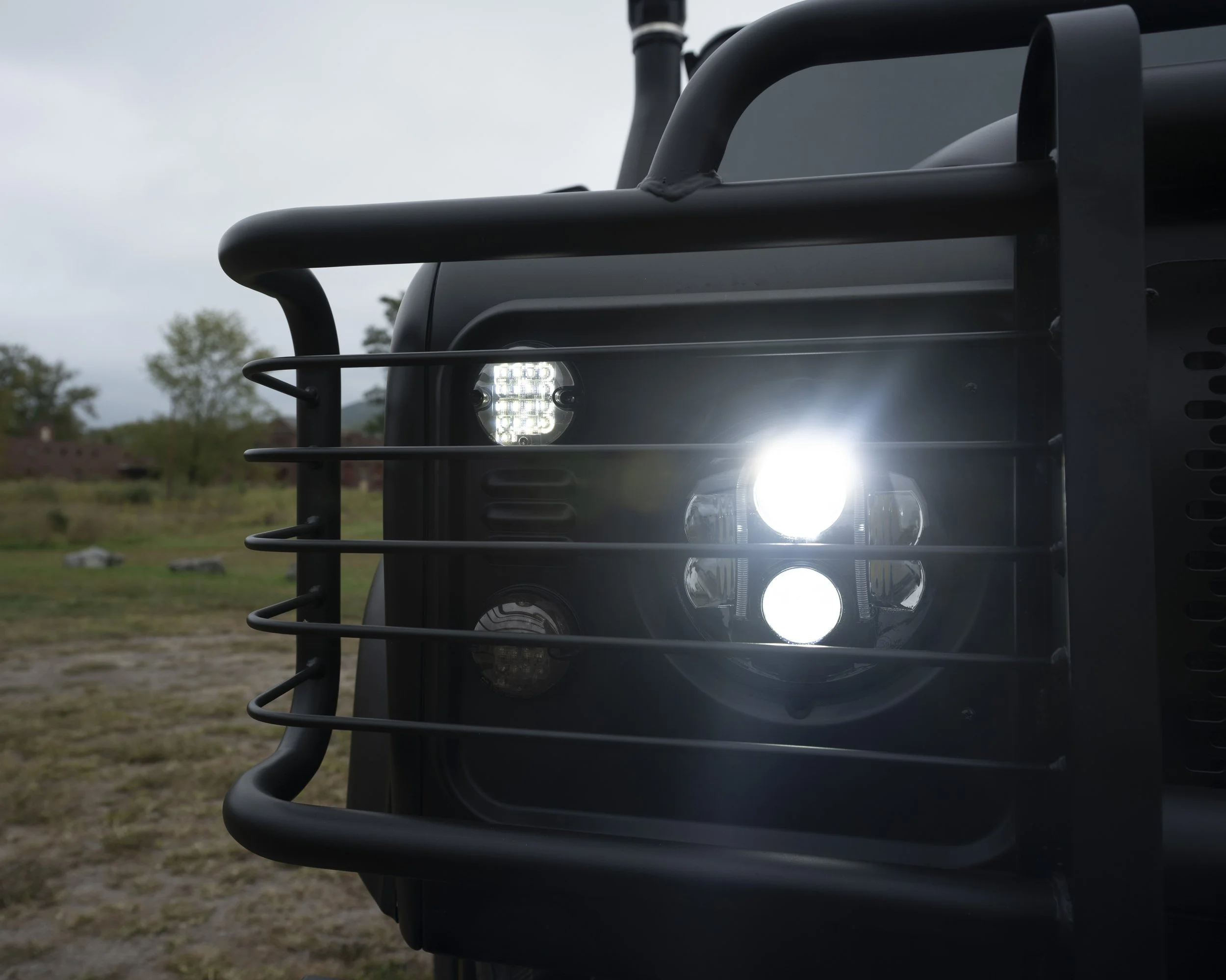 Close-up of the front of a black off-road or utility vehicle with a metal grille, featuring round headlights and multiple smaller LED lights, set outdoors on a cloudy day.