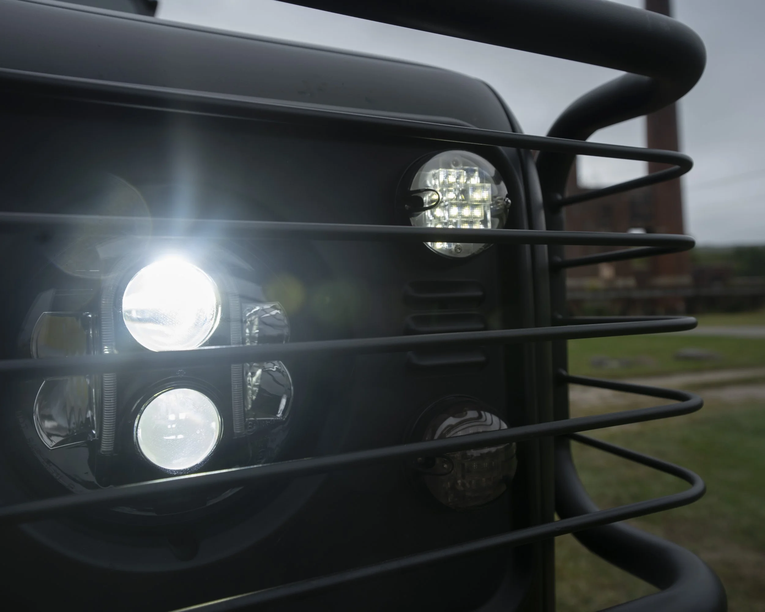 Close-up of a vehicle's headlight with a protective grill, with the light turned on and the sky visible in the background.