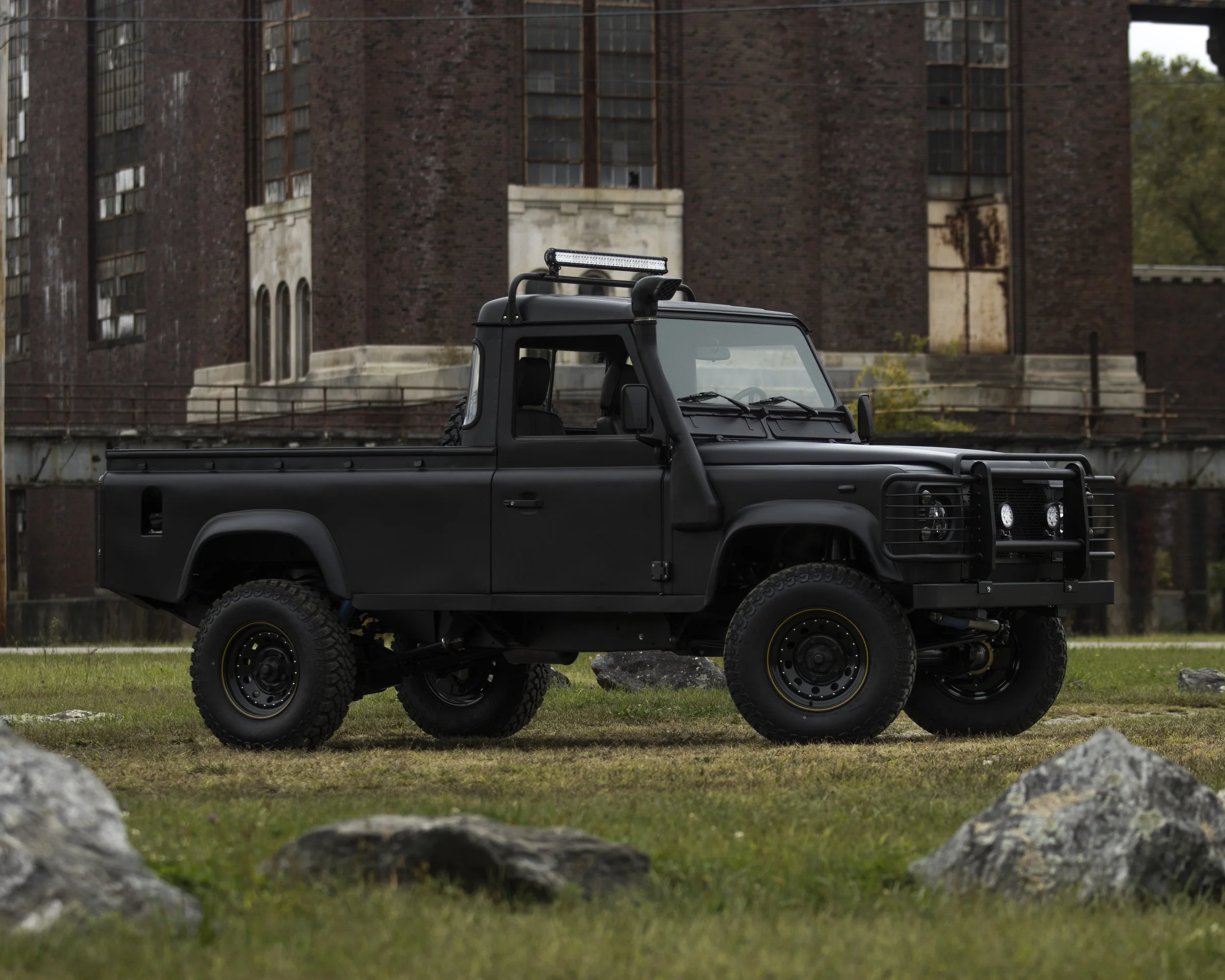 A black off-road pickup truck parked on grass with rocks in the foreground and an old brick building in the background.