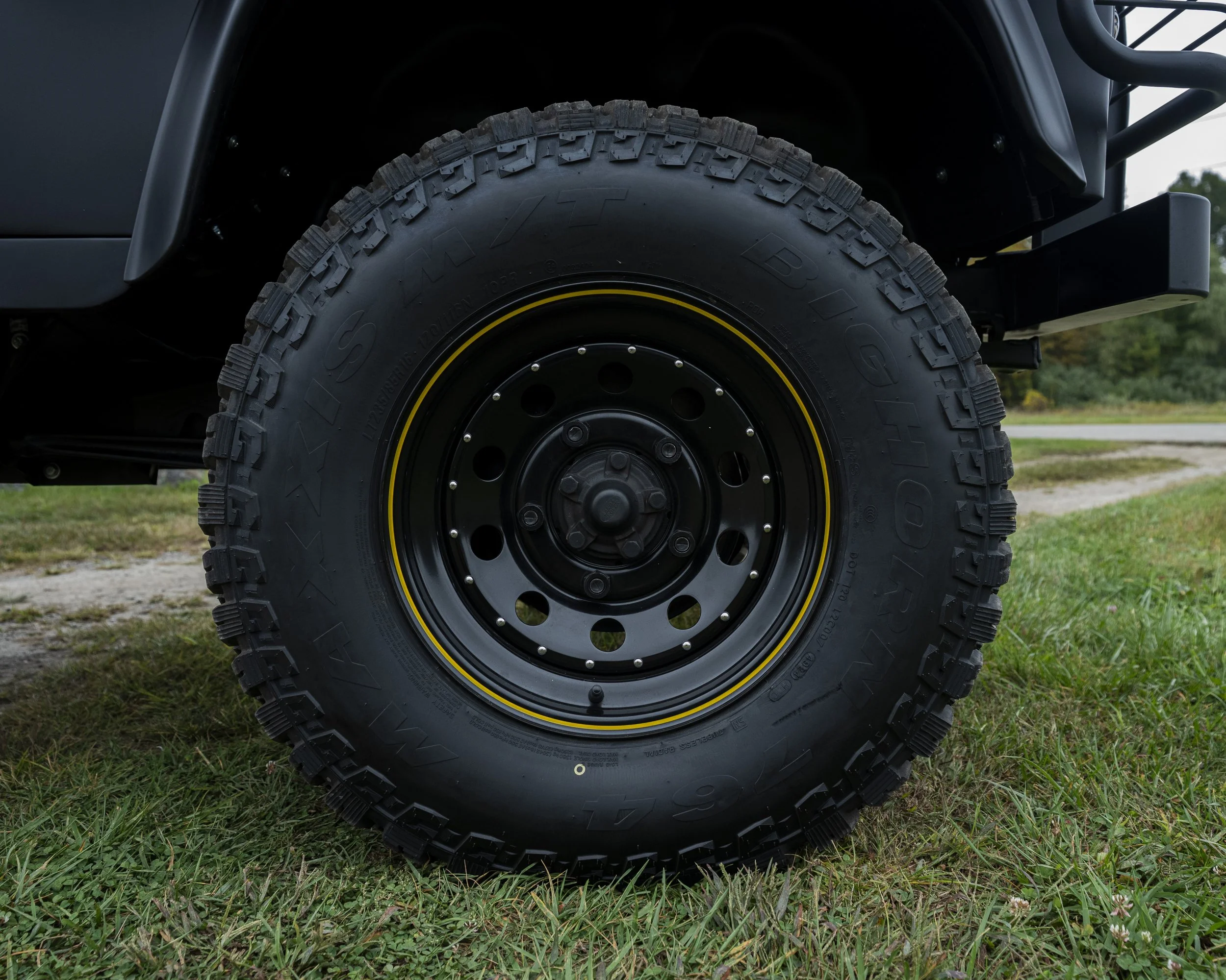 Close-up of a large off-road tire mounted on a black rim, with a grassy area and dirt path in the background.