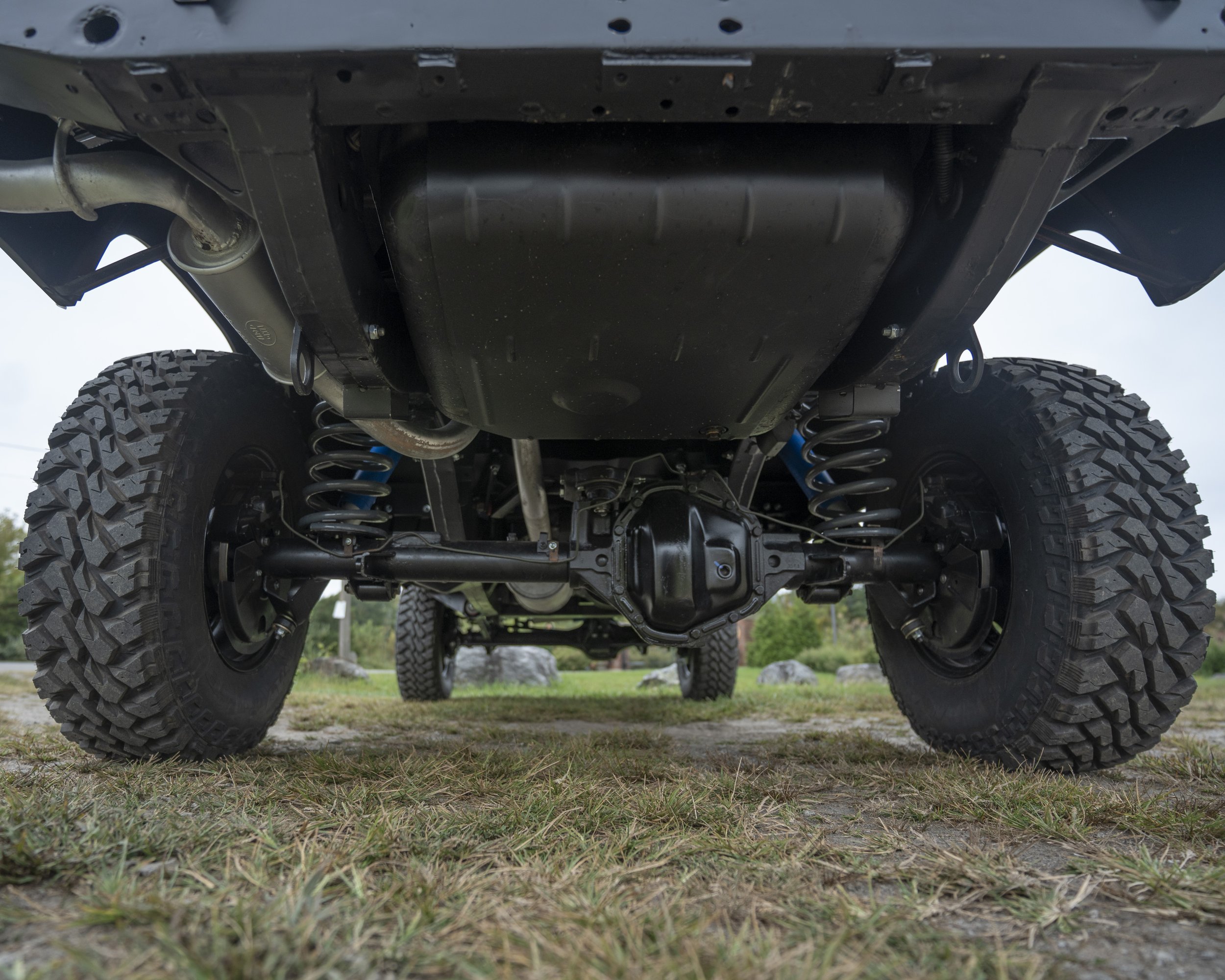 Underneath view of a lifted off-road vehicle showing suspension, differential, and large rugged tires on grassy ground.