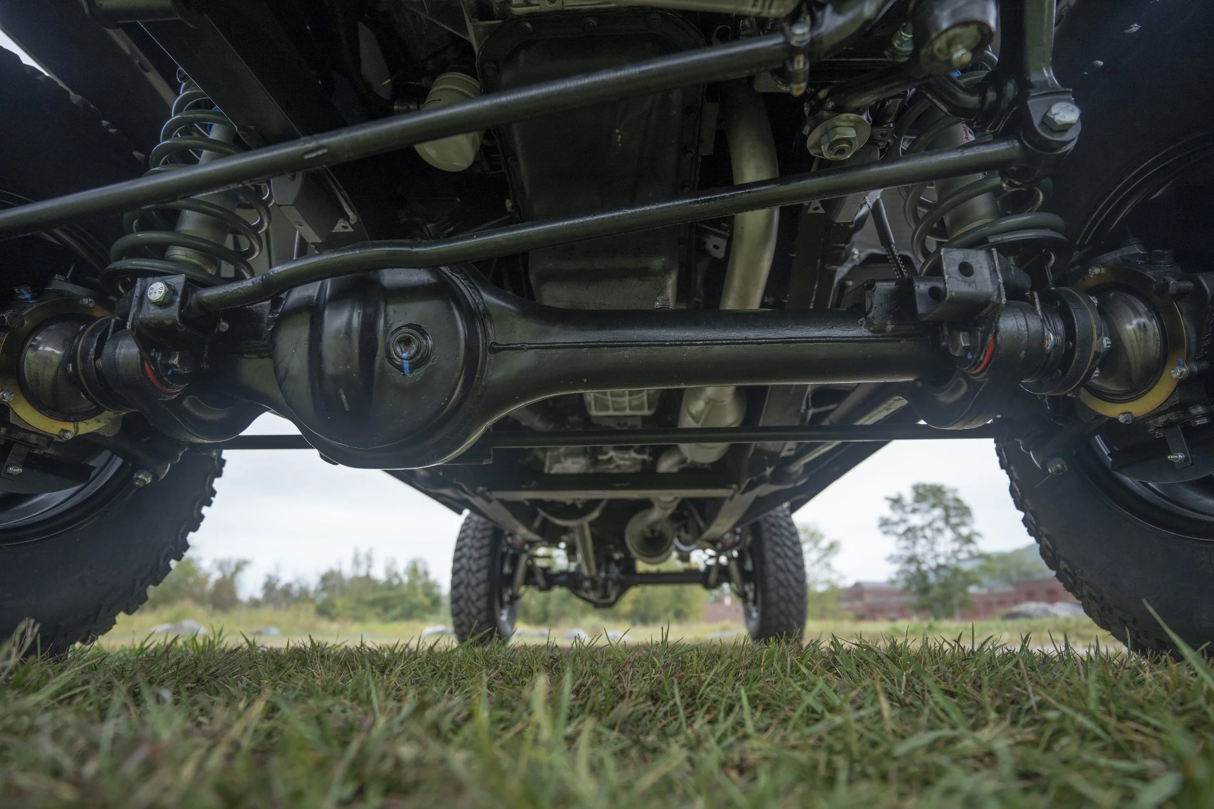 Underneath view of a vehicle showing suspension components, axle, and tires on grass with trees and a building in the background.