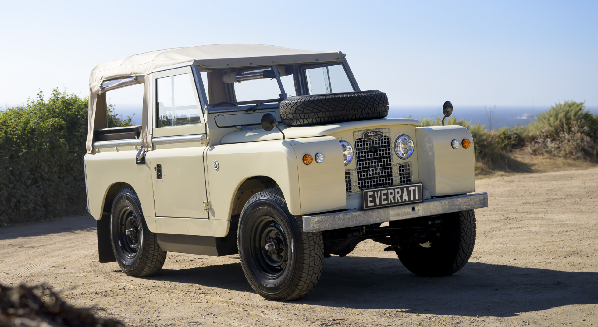 A vintage beige Land Rover parked on a dirt road with bushes and the ocean in the background.