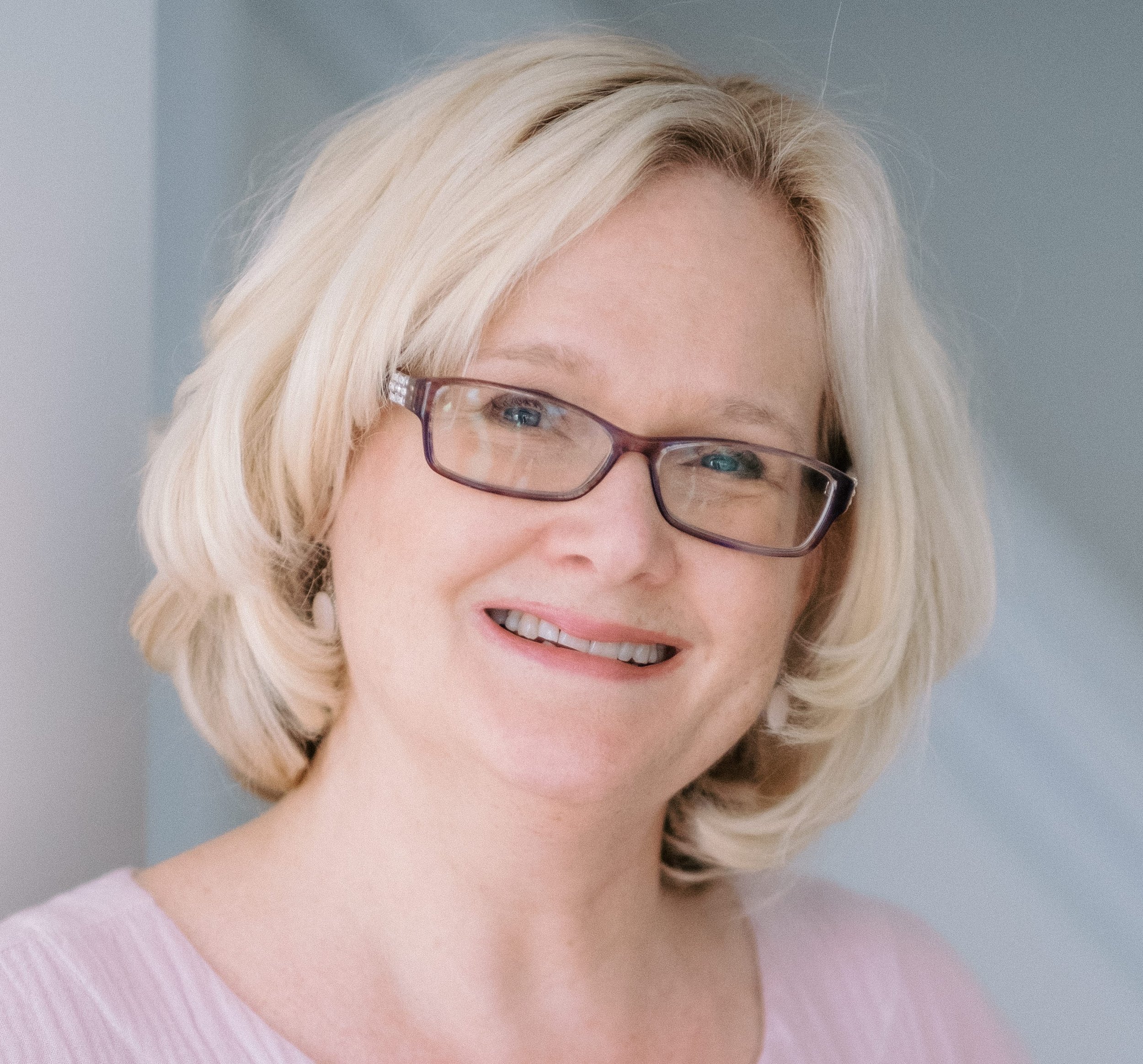 A smiling middle-aged woman with blonde hair, wearing glasses, and a light pink top.