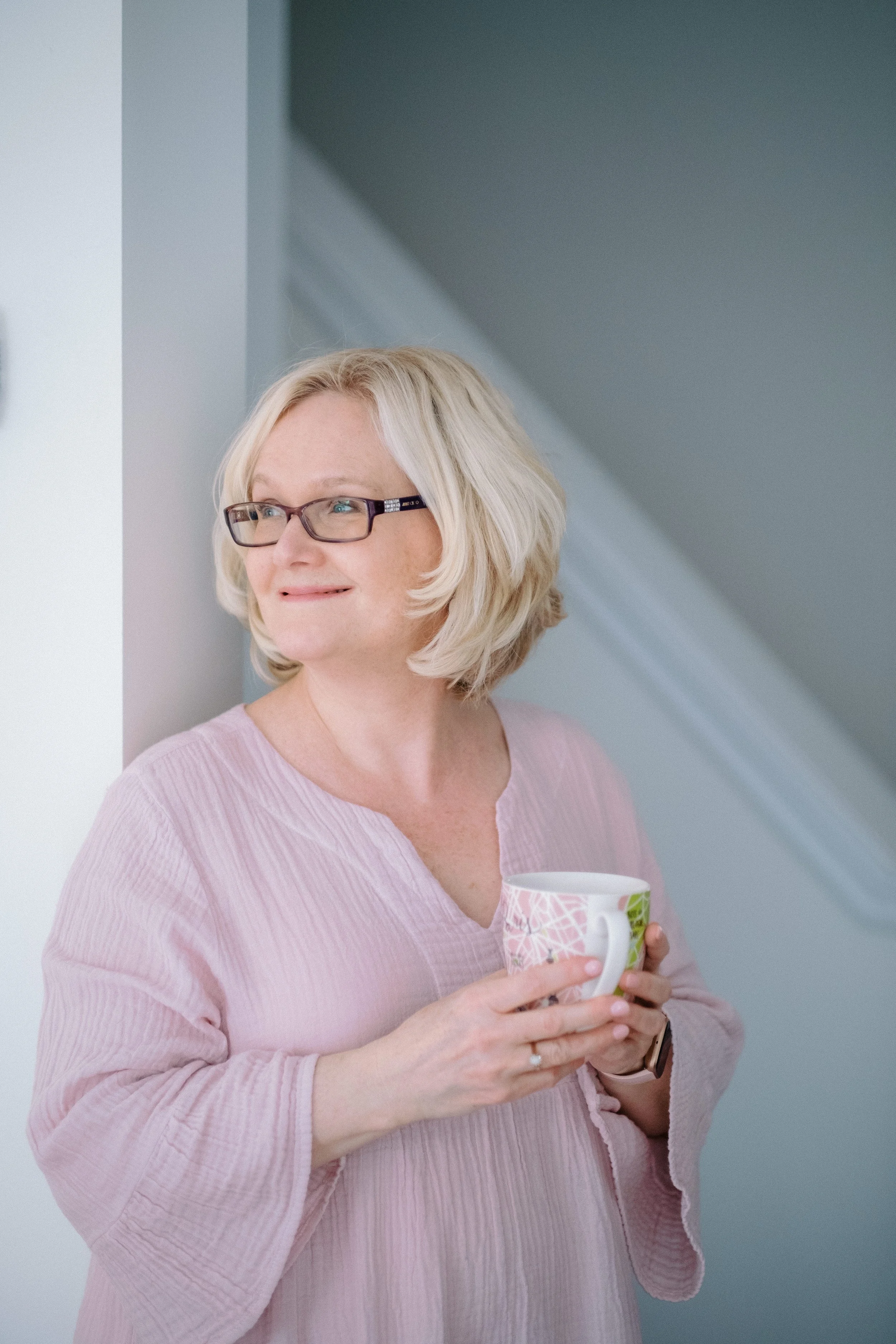 A middle-aged woman with blonde hair, glasses, wearing a pink top, holding a colorful mug, standing indoors.