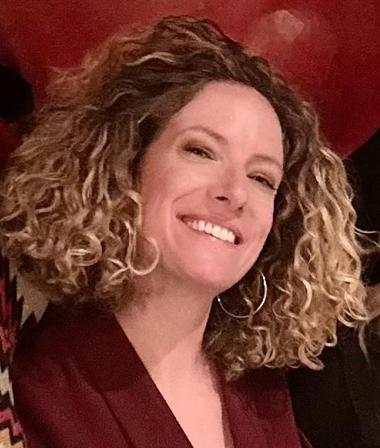 A woman with curly blond hair smiling in front of a red background.