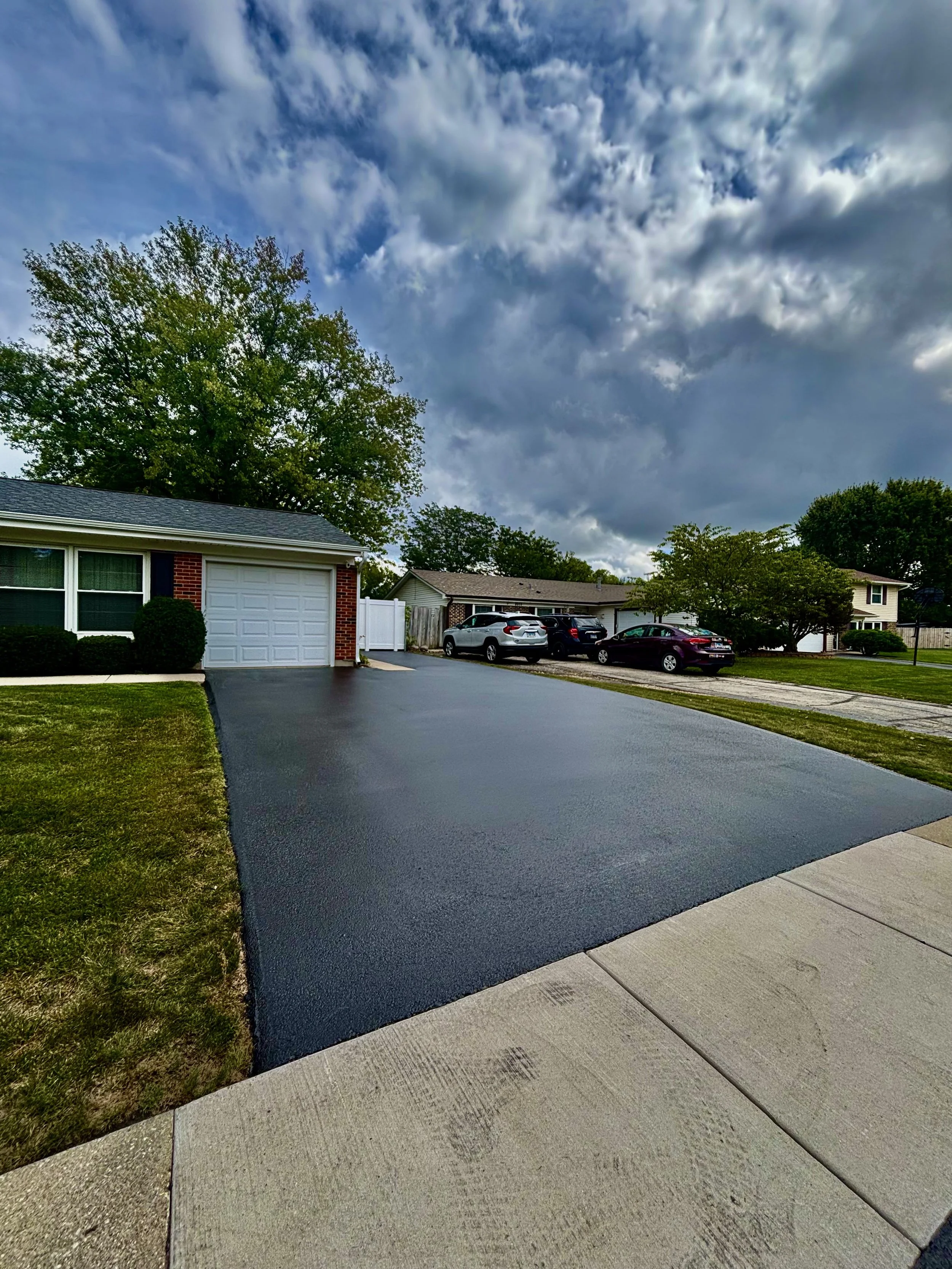 freshly sealed asphalt driveway leading to a white house, flanked by green bushes and trees on both sides.