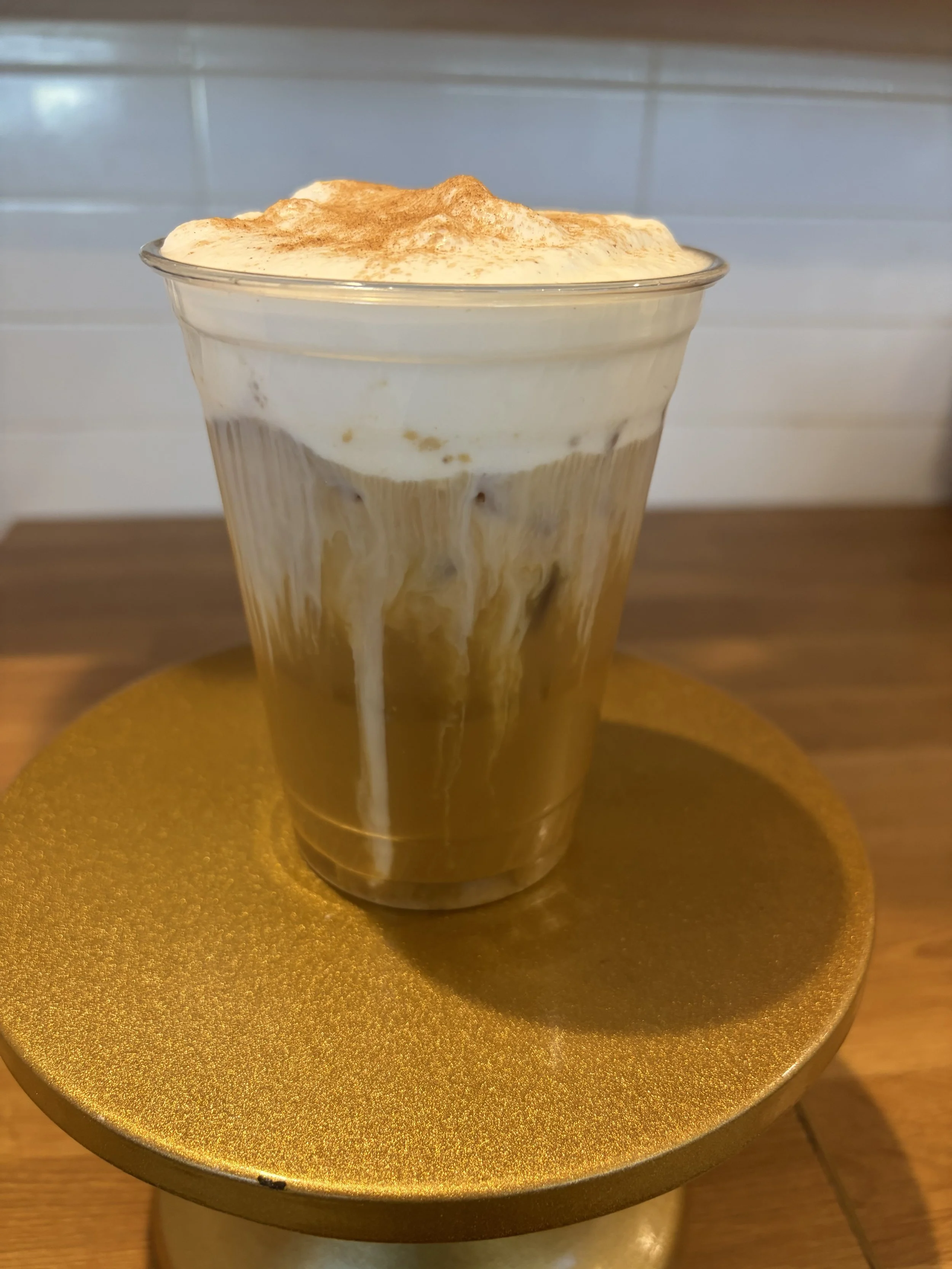An iced latte with a vanilla white cold foam on a gold stand with a white tile background