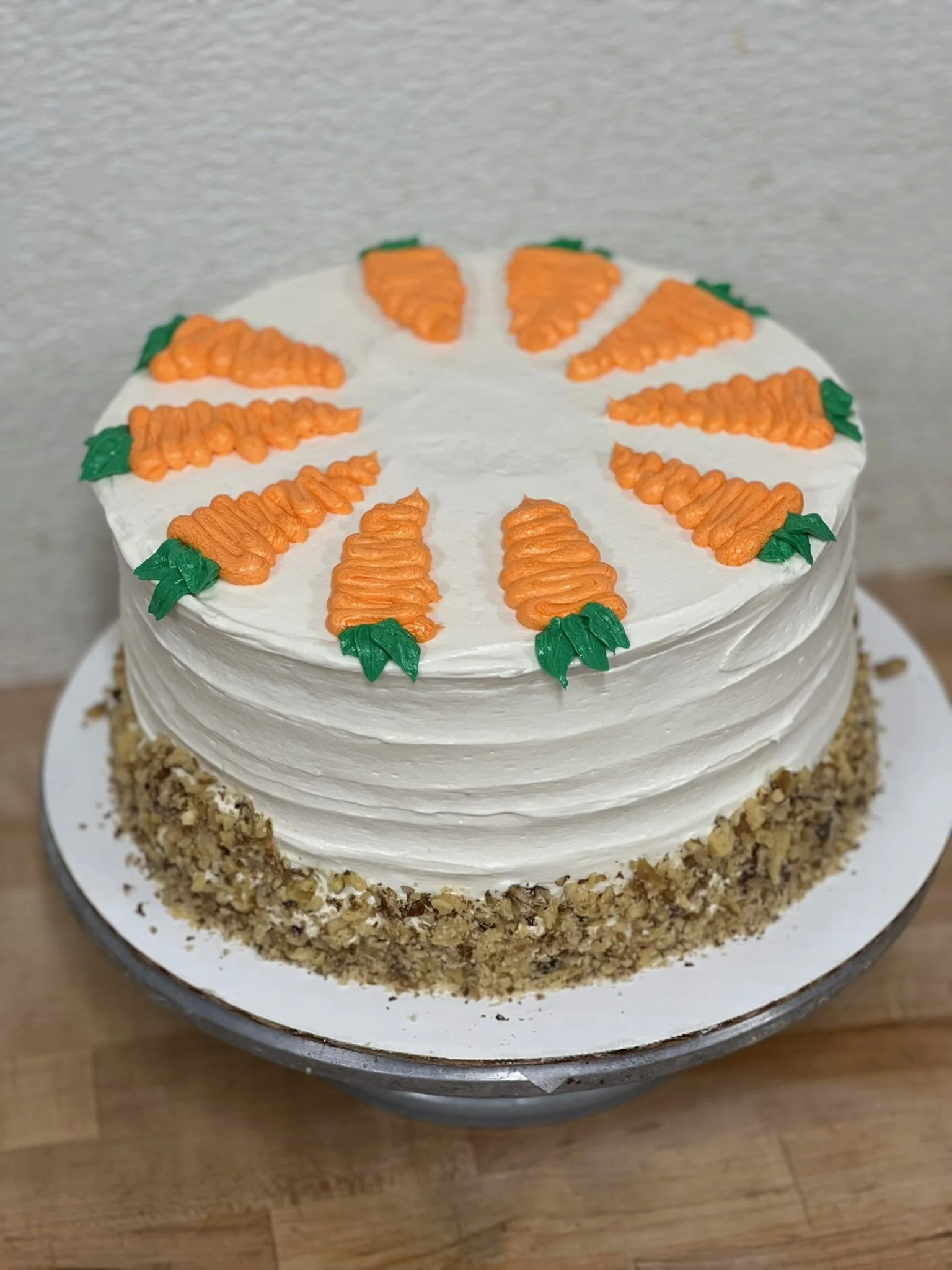 A carrot cake covered in white icing with a lower band of crushed walnuts decorated with orange and green frosting carrots